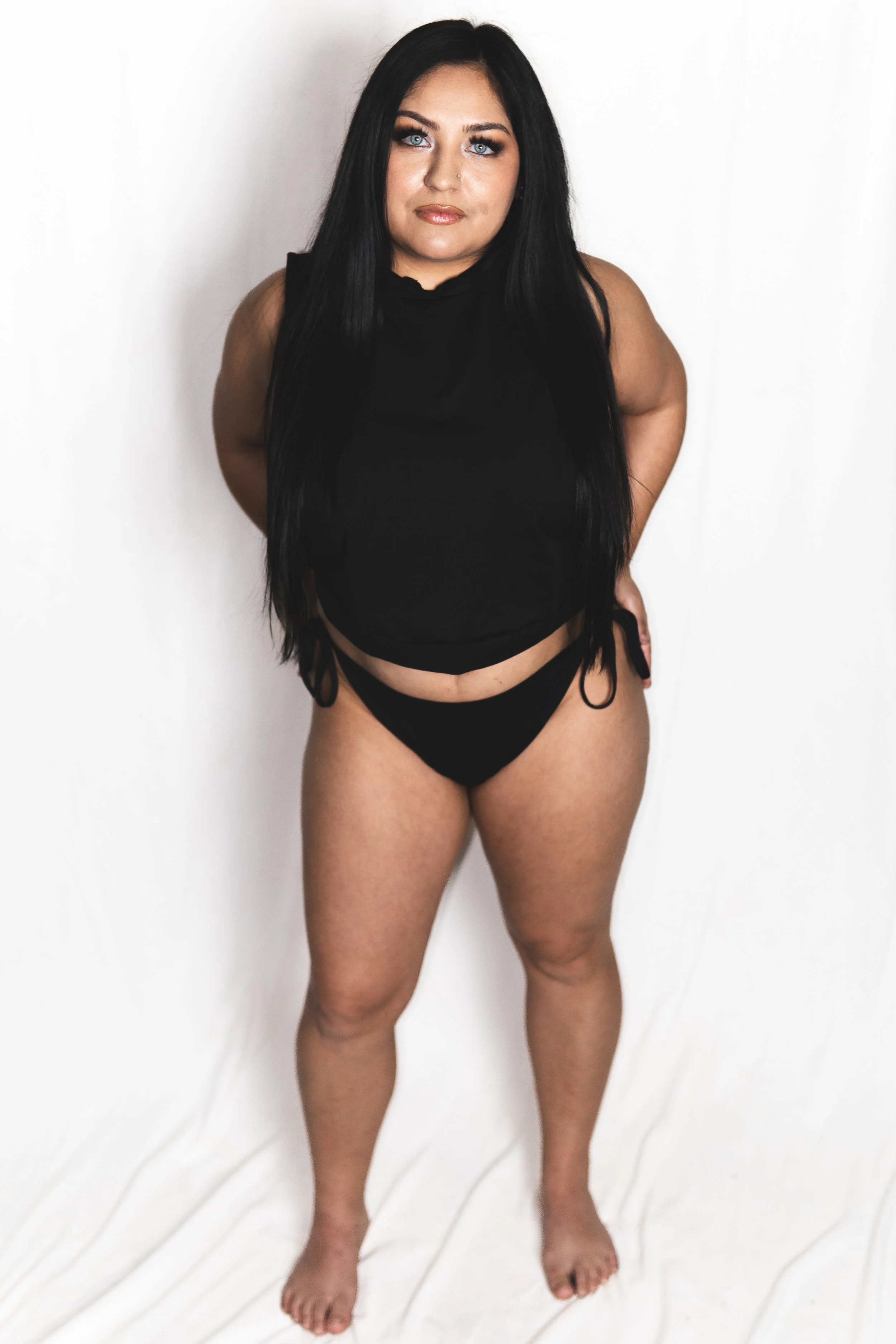 A woman with long black hair, blue eyes, and light skin stands against a plain white background. She is wearing a black sleeveless top and black underwear with side ties, standing with her hands behind her back.