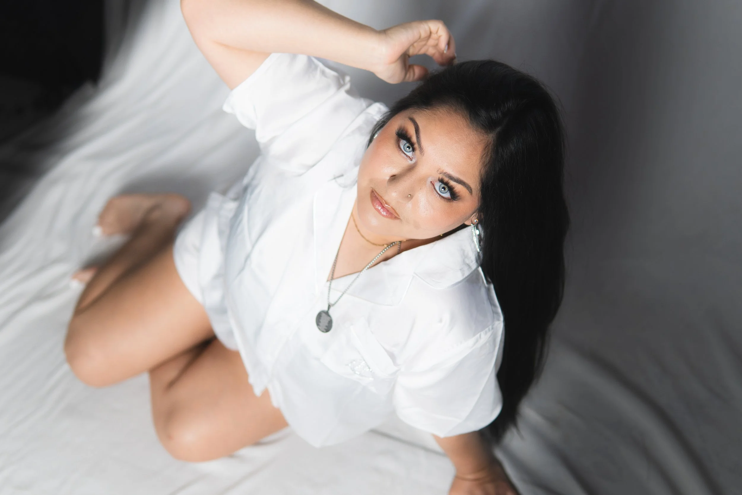 A woman with blue eyes, dark hair, and a nose piercing is lying on a bed wearing a white shirt. She is looking directly at the camera and has one hand roughing her hair.