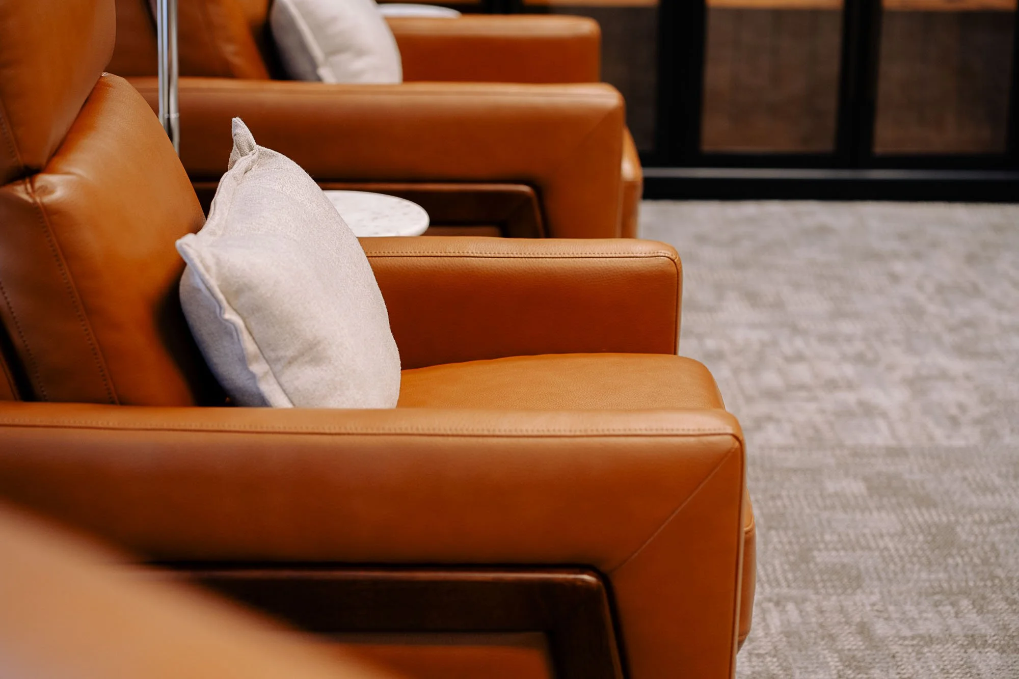 Row of tan leather chairs with white cushions in a waiting room or lounge area, with a carpeted floor.