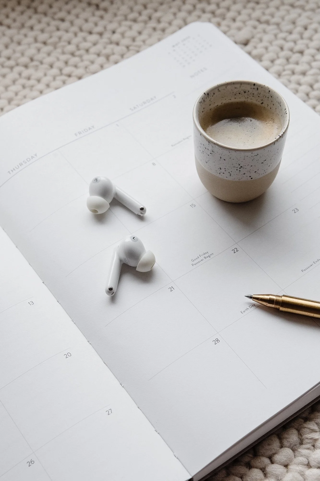 A calendar, a coffee cup, wireless earbuds, a gold pen, and a notebook on a textured surface.