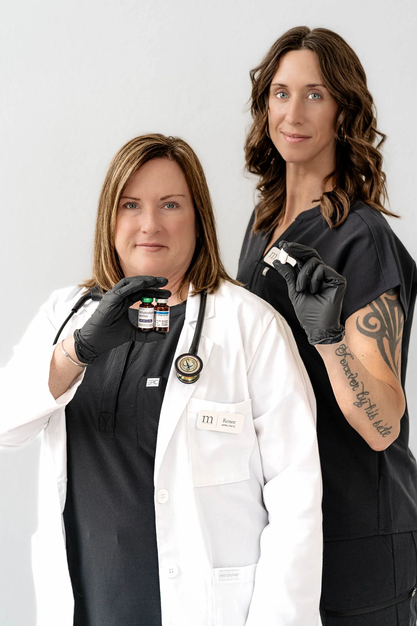 Two women, one dressed as a doctor and the other as a tattoo artist, are posing together. The woman in the white coat is holding a small vial labeled 'ethicobal' and is wearing a stethoscope. The woman in dark scrubs has tattoos on her arm and is holding a small object, possibly a vial or medication.