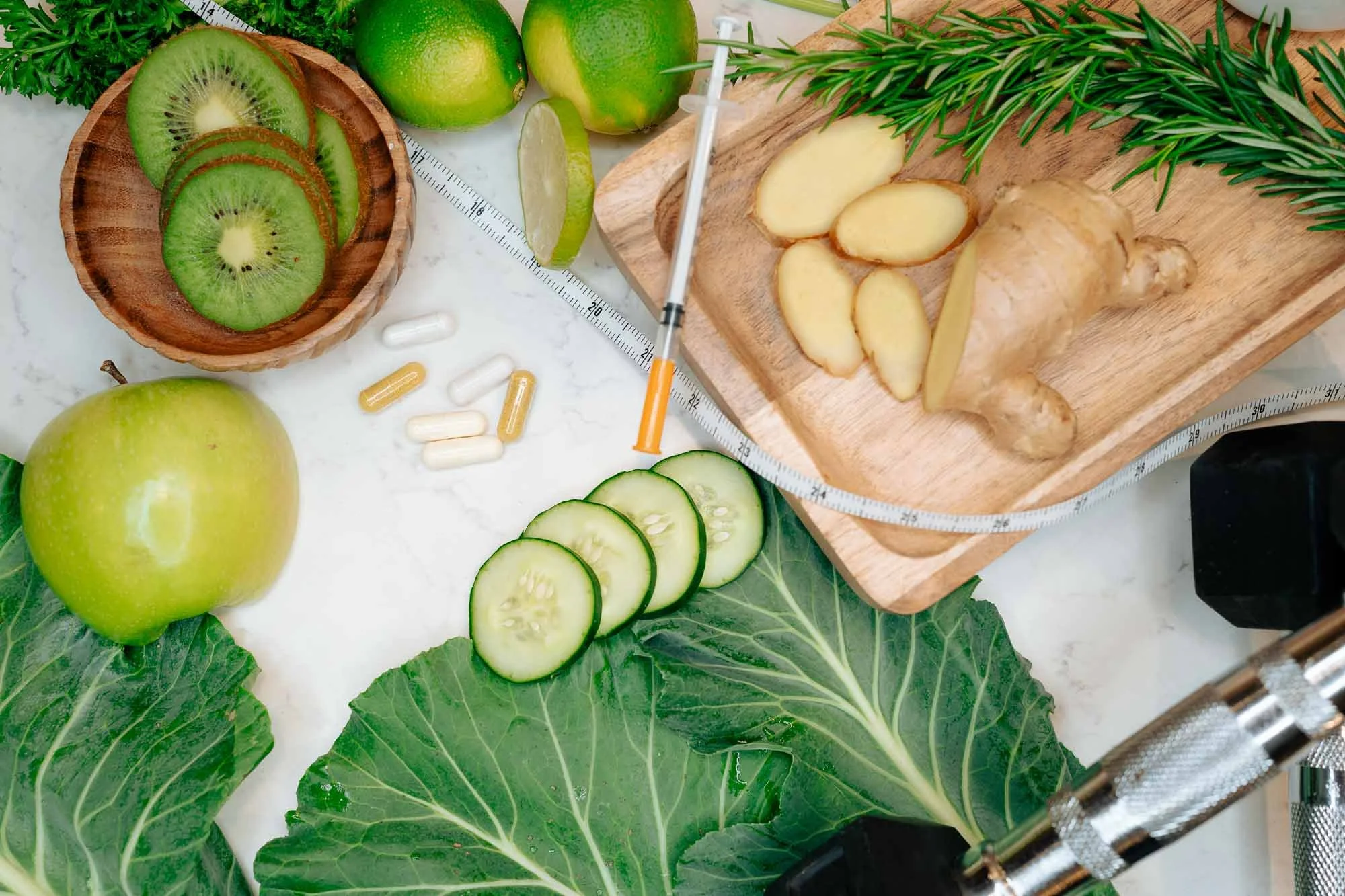 Assorted fresh herbs, fruits, and vegetables on a kitchen countertop, including sliced kiwi, green apples, limes, cucumbers, ginger, and sprigs of rosemary and leafy greens, with a syringe among the ingredients.