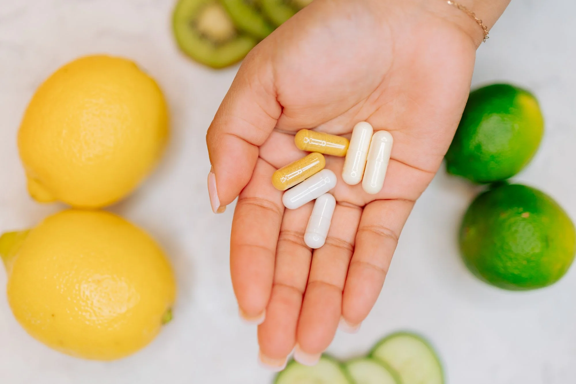 A person's hand holding qeuantity of capsules with yellow and white color, surrounding citrus fruits, including lemons and limes, with sliced cucumber in the background.