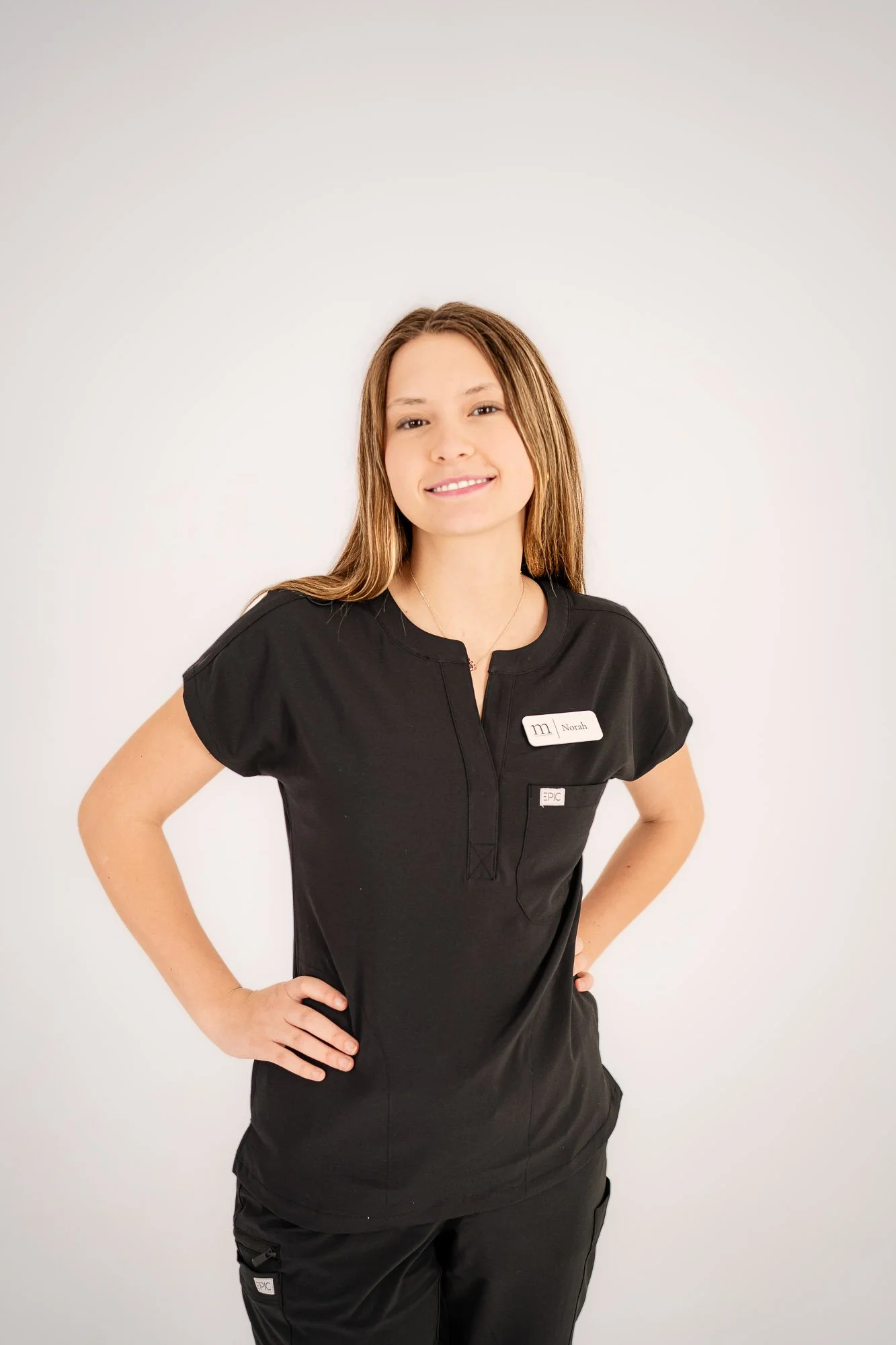 A woman wearing a black uniform with a name tag that reads 'Noah' and standing against a plain light-colored background.