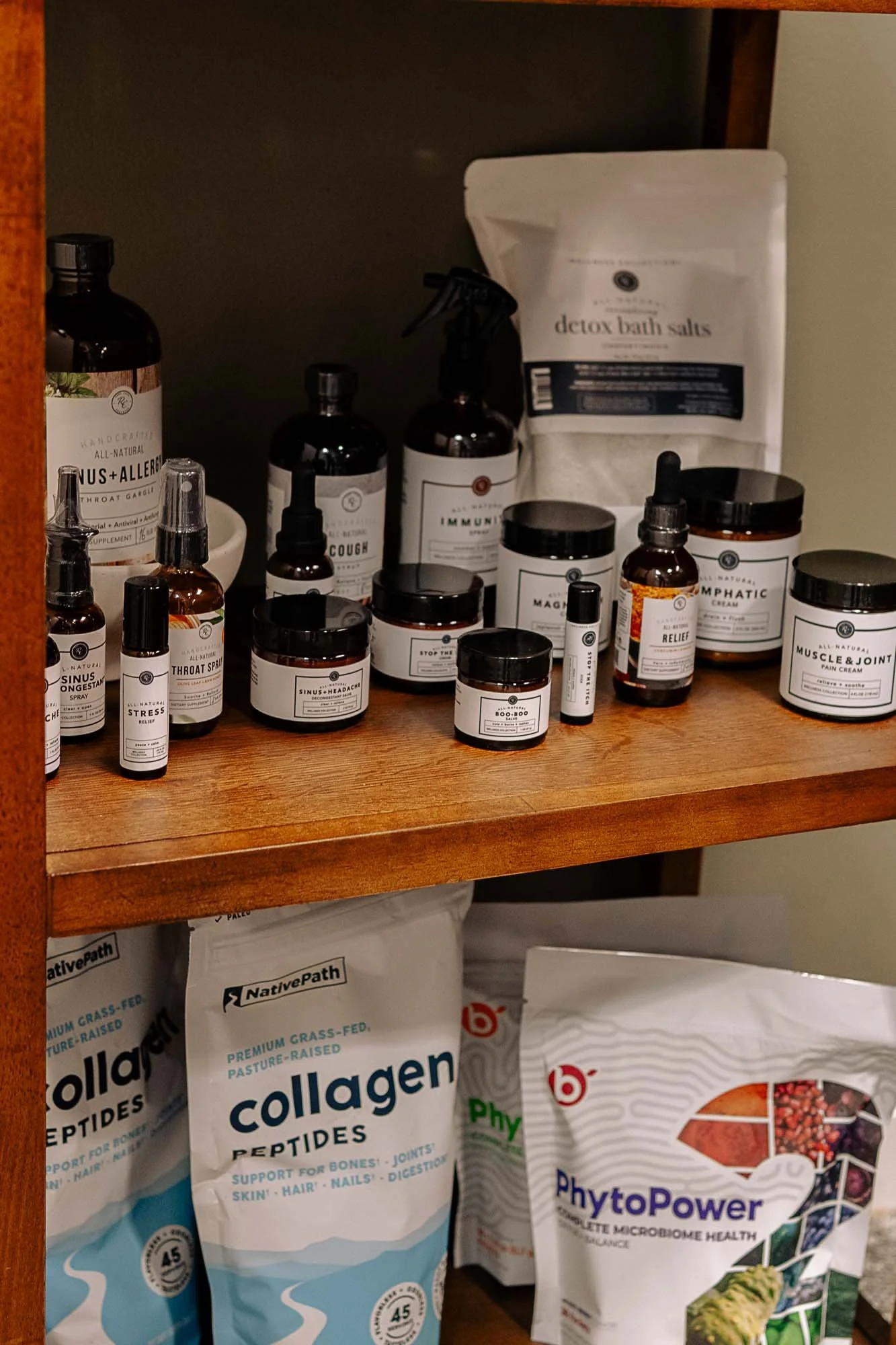 A wooden shelf holding various health and wellness products including bottles of supplements, a bag of detox bath salts, and bags of collagen peptides on the lower shelf.