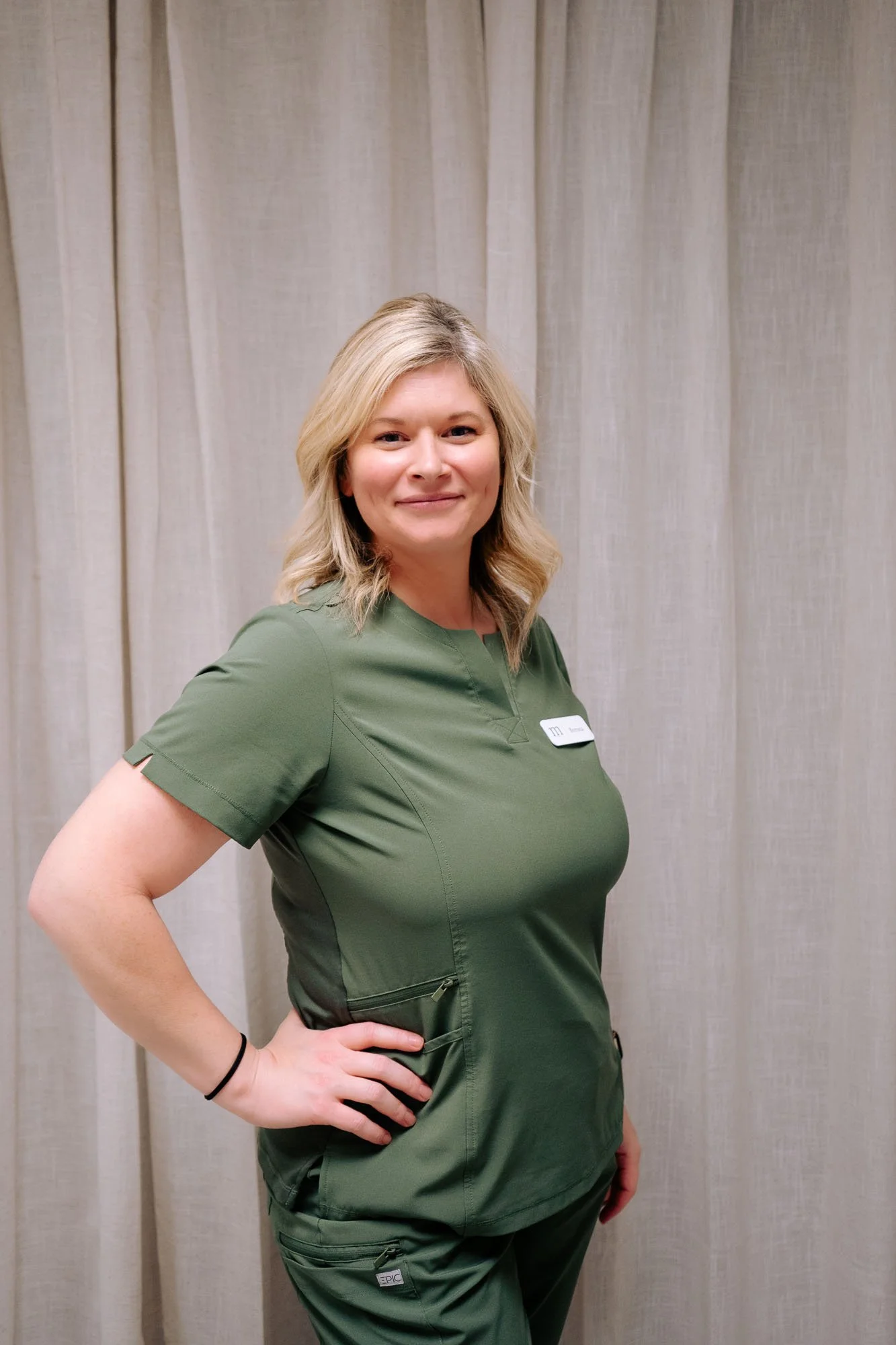 A smiling middle-aged woman with blonde hair, glasses, and hoop earrings wearing a black uniform with a name tag, standing against a plain white background.
