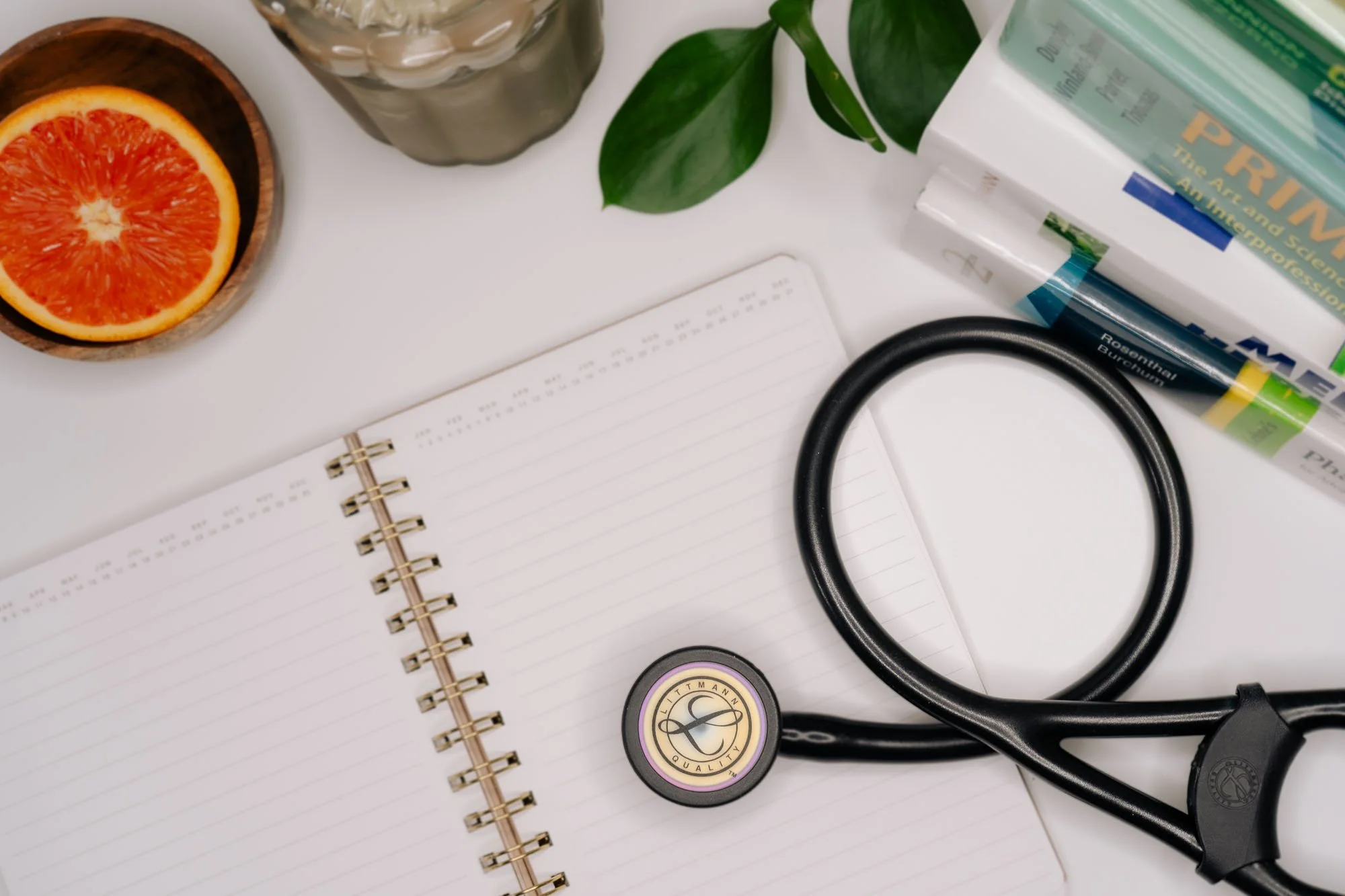 A flat lay of medical and office supplies on a white surface, including a stethoscope, a spiral notebook, a halved orange in a wooden bowl, a glass of water, green leaves, and boxes of medical gloves.