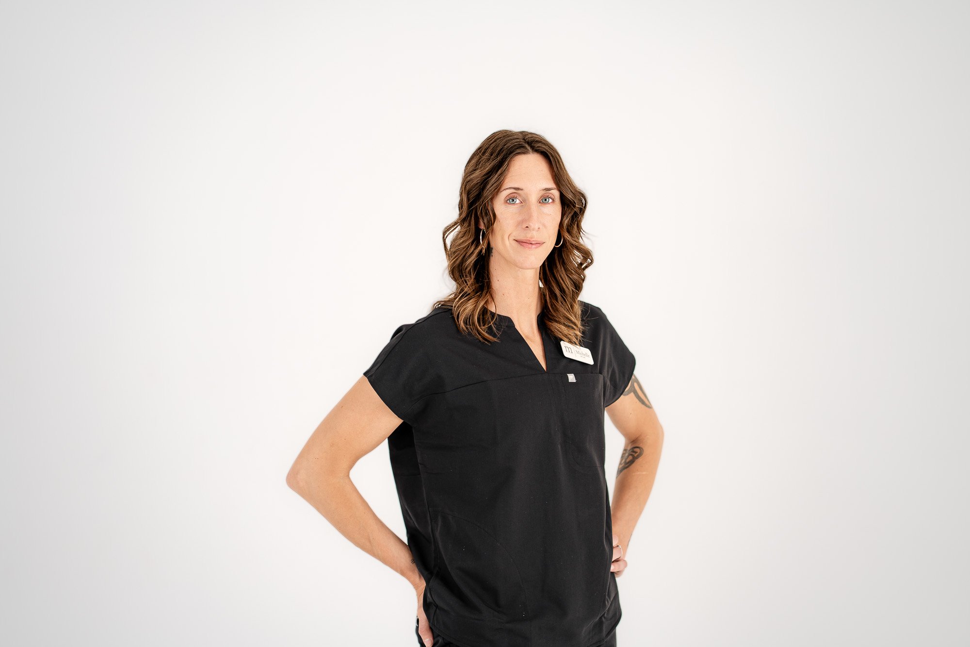 A woman with wavy brown hair and blue eyes wearing a black uniform with a name tag, standing with her hand on her hip against a plain white background.