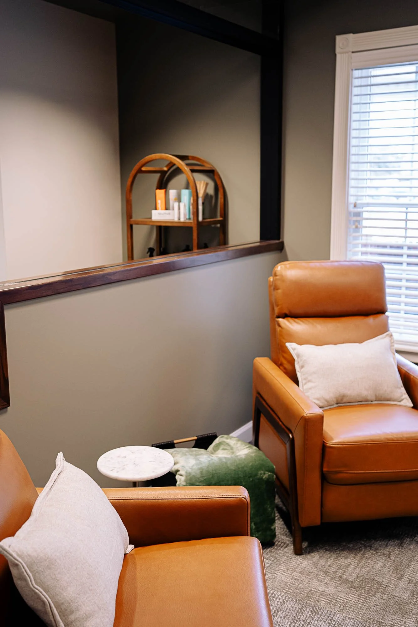A cozy sitting area with tan leather chairs, a small white marble side table, a cream pillow, and a green blanket. In the background, a large window with white blinds and a wall with a mirror and a wooden shelf with books and decor are visible.