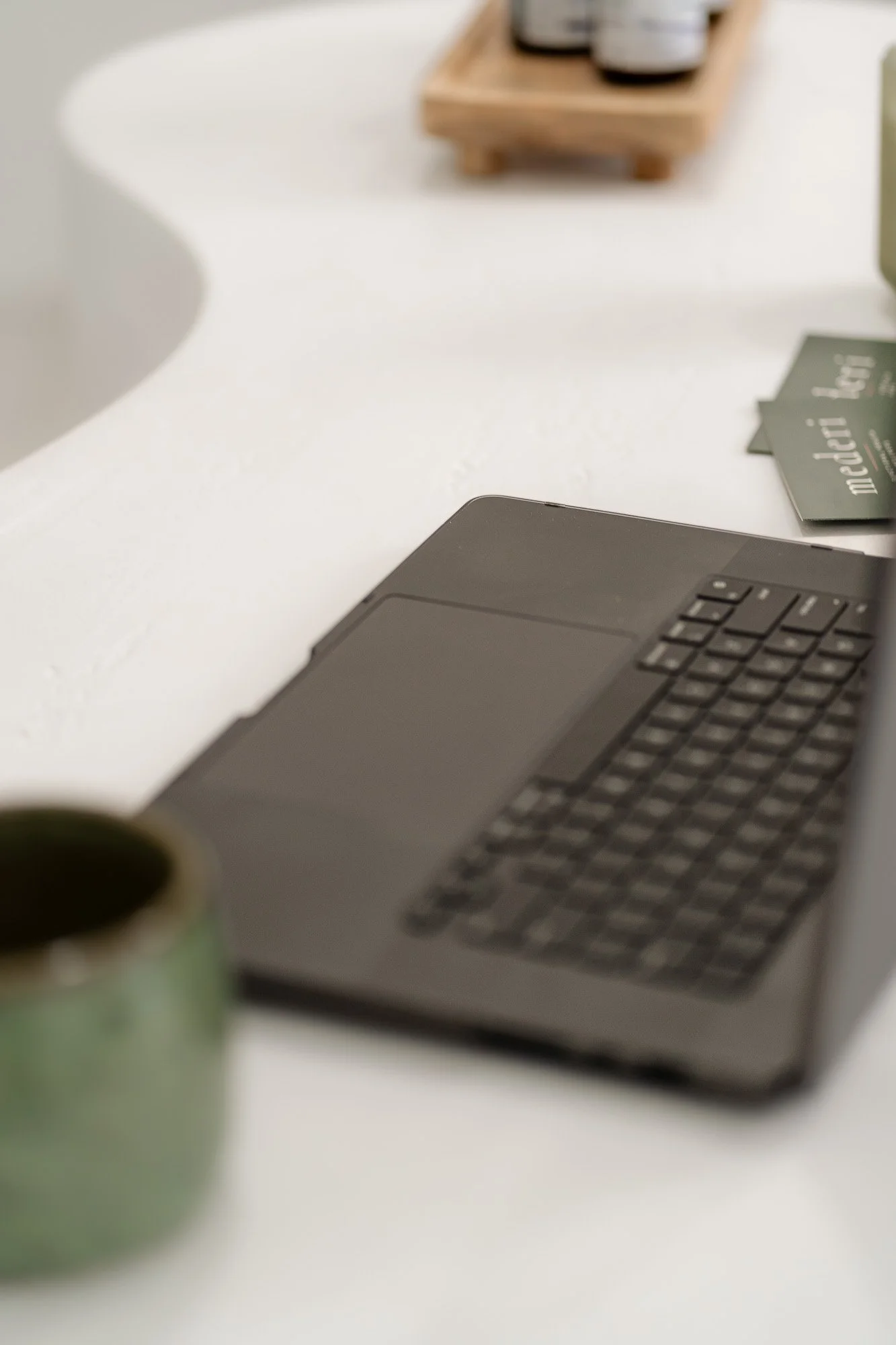 Close-up of a laptop computer on a white desk, with a blurred green mug in the foreground and a small wooden tray with bottles and containers in the background.