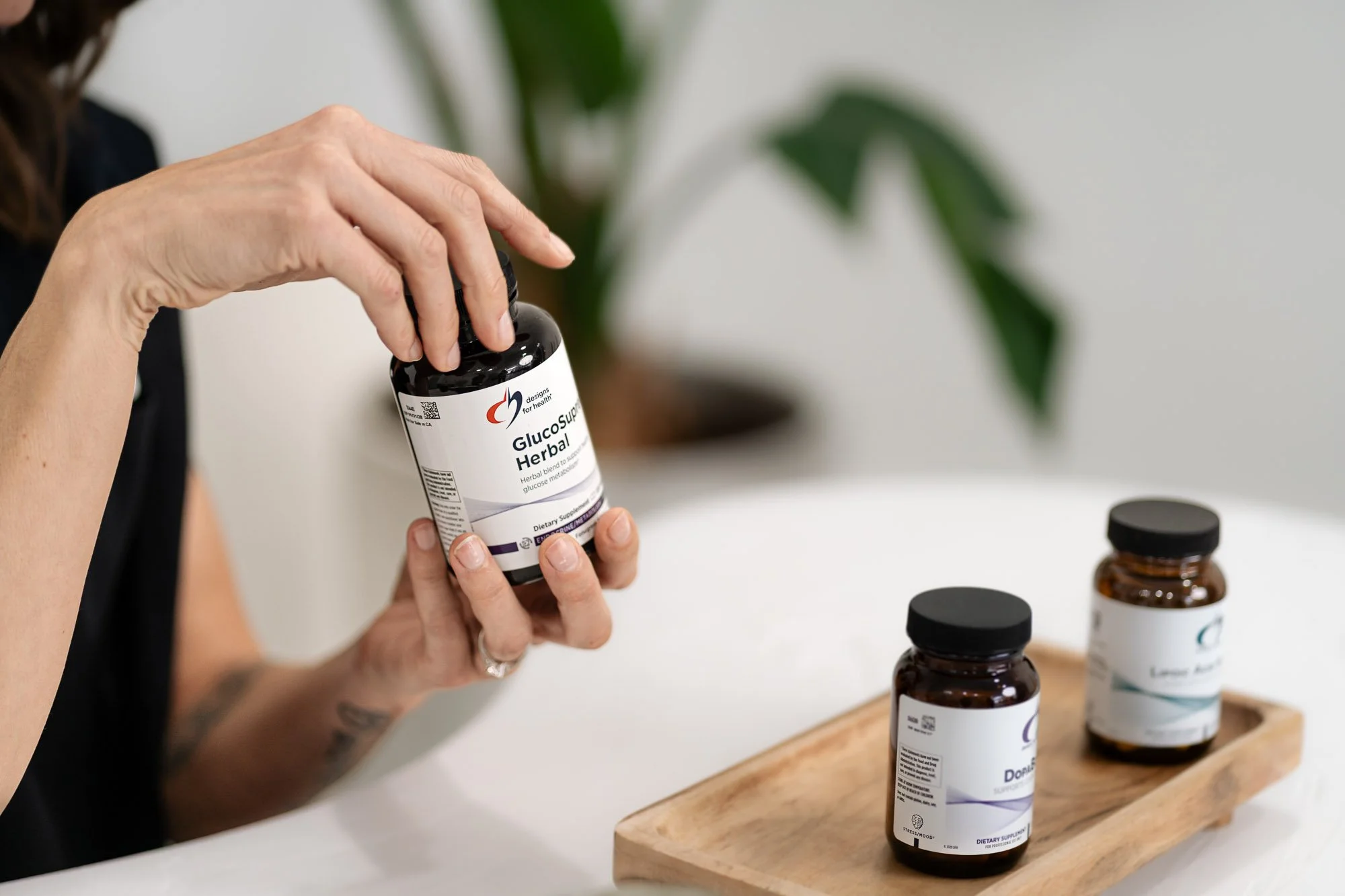 A person holding a bottle labeled 'Glucose Support Herbal' supplement, with other supplement bottles on a wooden tray nearby, in an indoor setting with a green plant in the background.