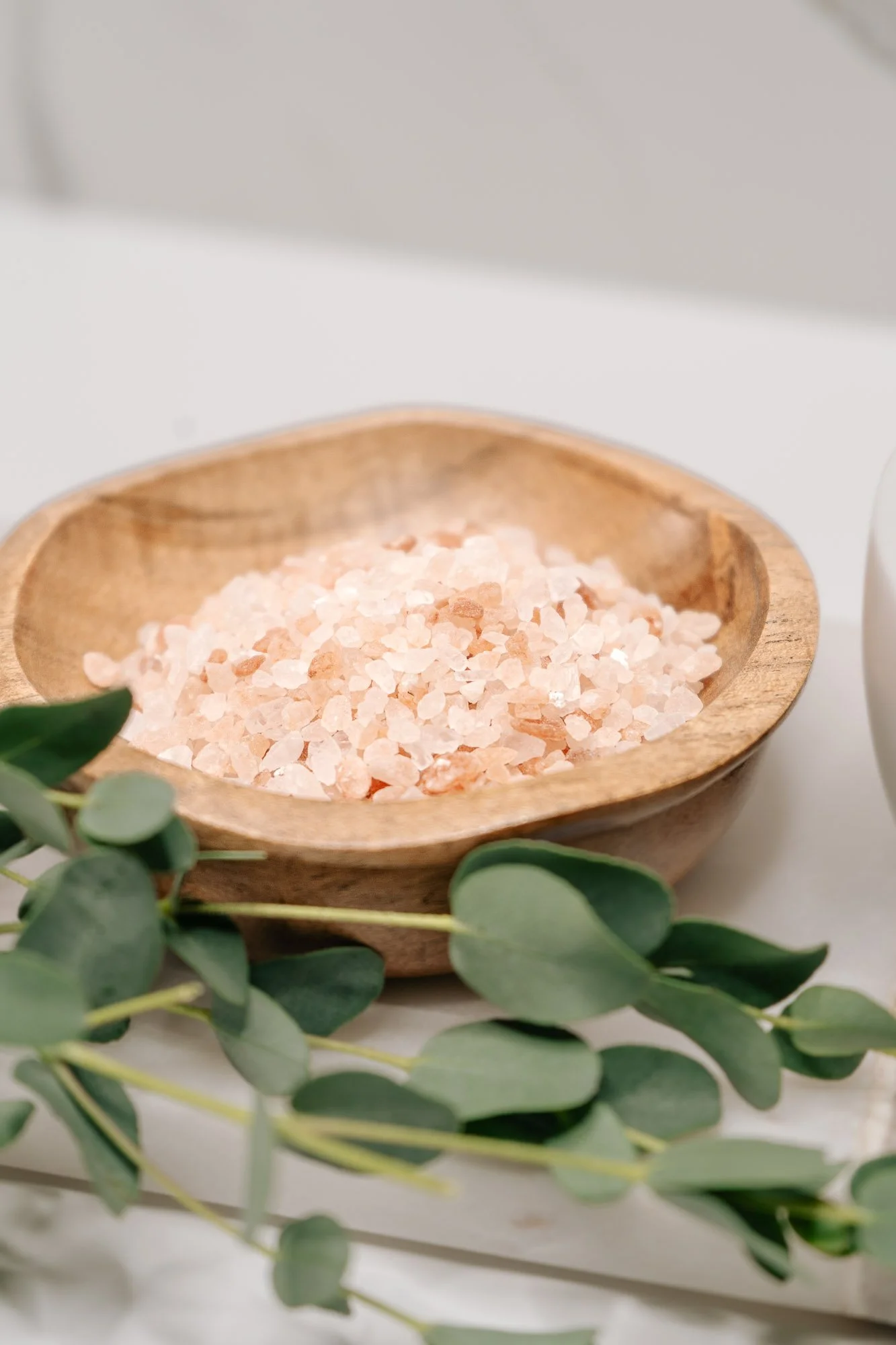 A wooden bowl filled with pink Himalayan salt and green eucalyptus leaves around it.
