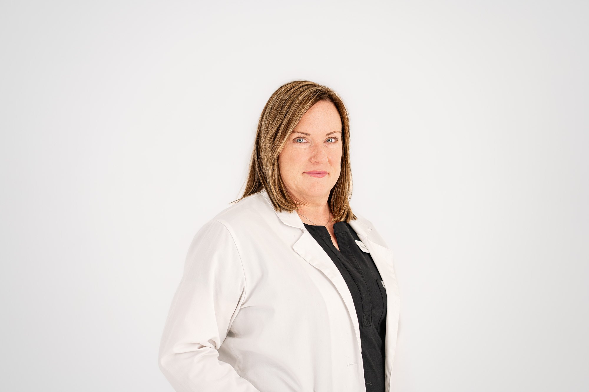 A woman with shoulder-length brown hair and blue eyes wearing a white lab coat over a black shirt, standing against a white background.