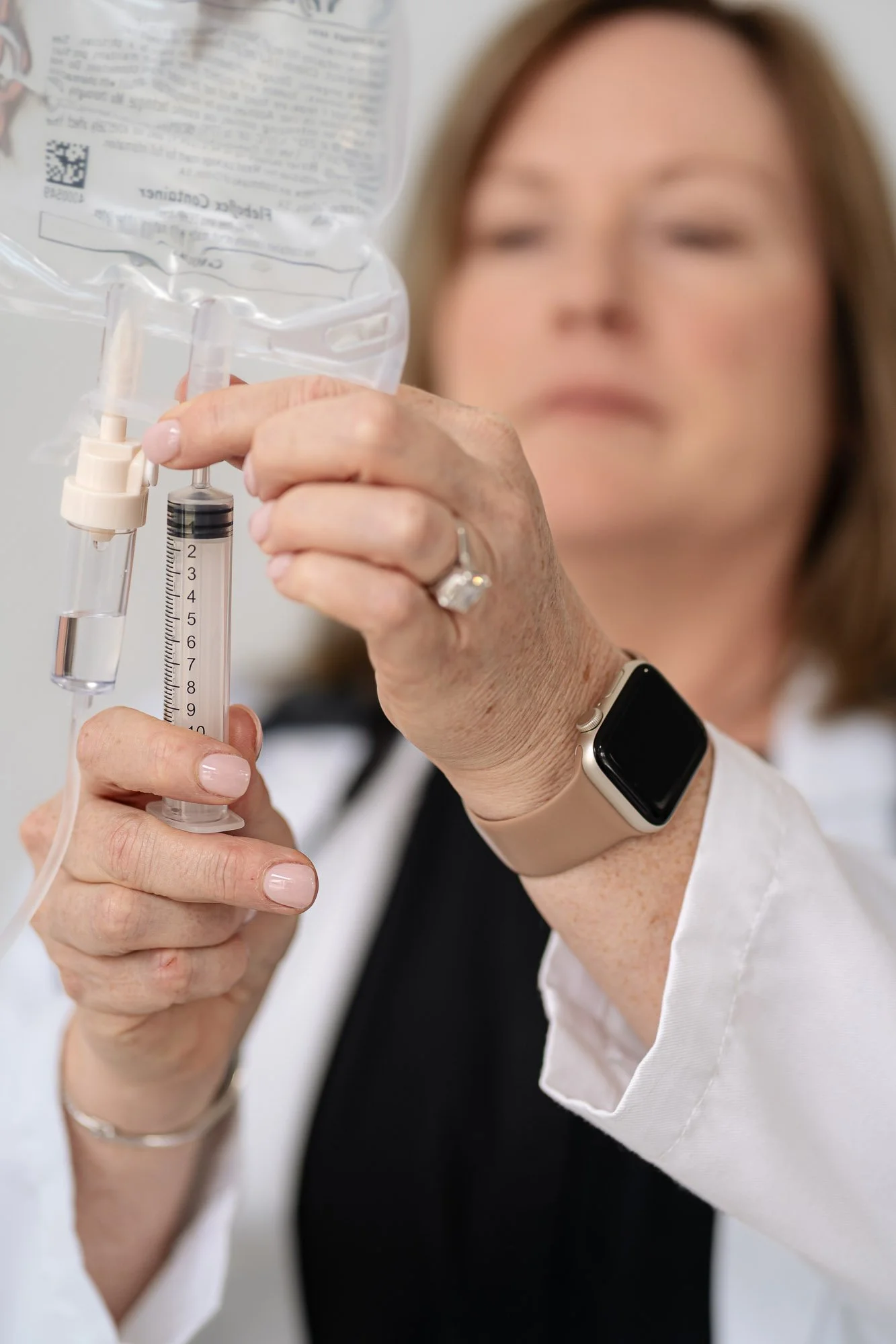 A woman preparing an injection with a syringe and a medication bag, wearing a smartwatch on her wrist and a white lab coat.