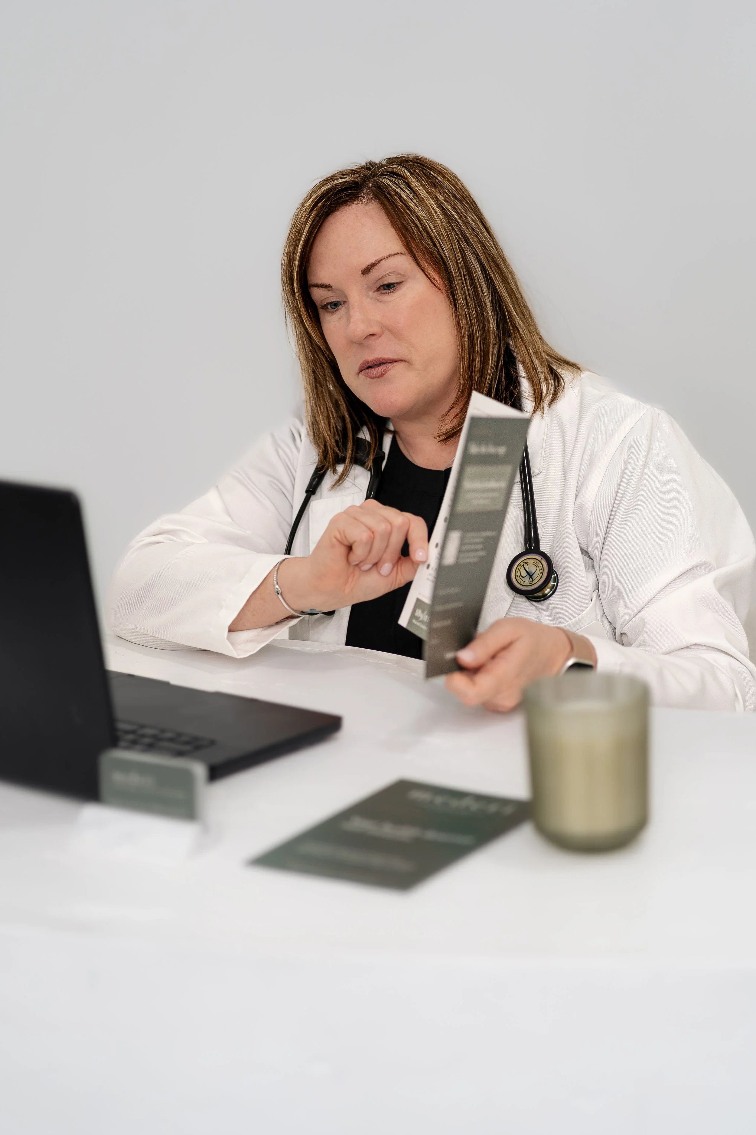 A woman in a white coat with a stethoscope on her neck, looking at a medical pamphlet in a clinical setting.