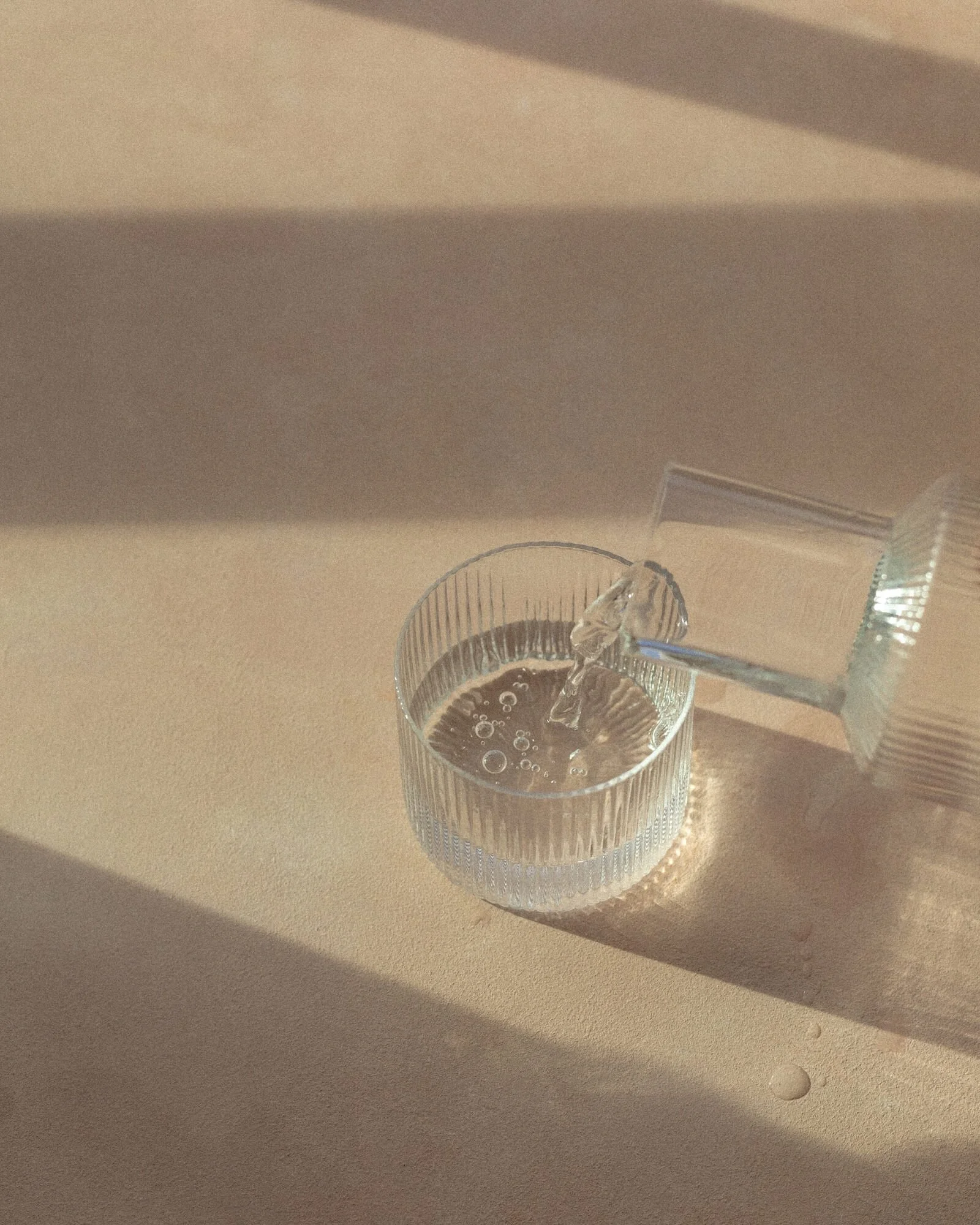 A glass of water with ice is being filled from a water pitcher on a beige countertop, with shadows cast by the glass and pitcher.
