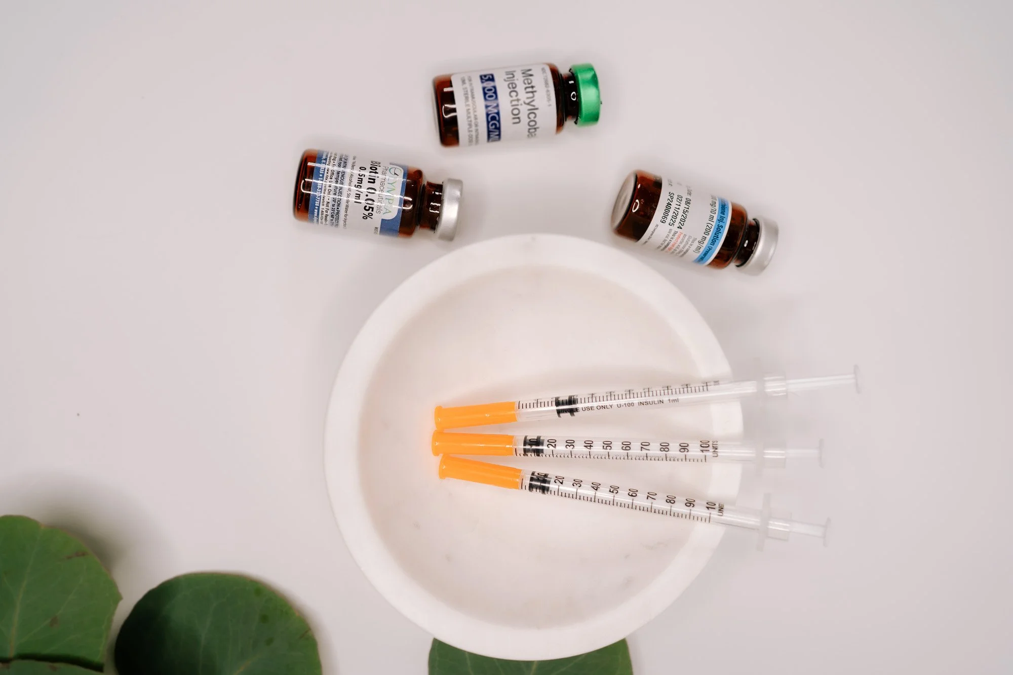 Three bottles of medication, several syringes with orange needles, and a small dish on a white surface with some green leaves at the bottom left corner.