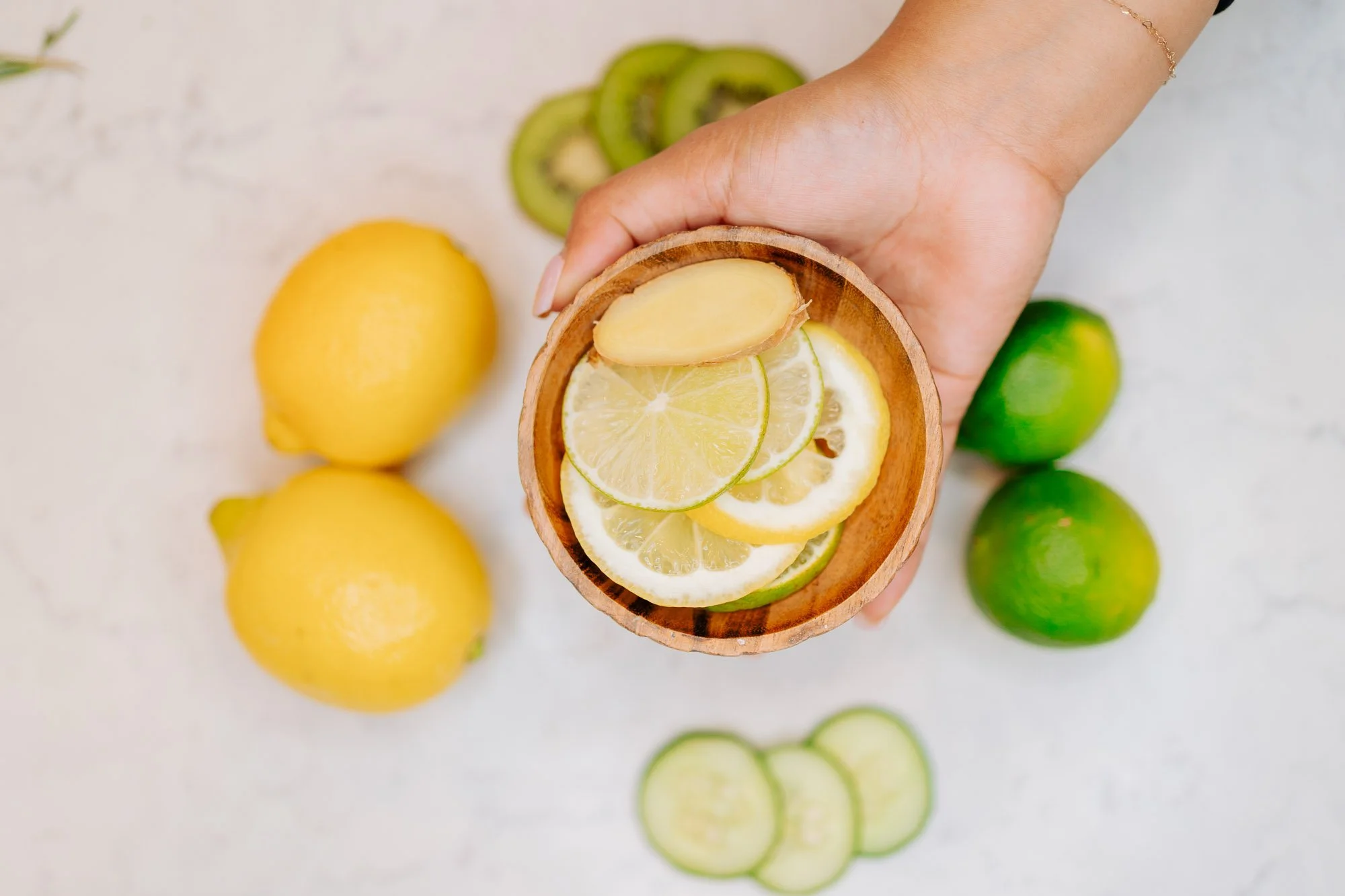 A person's hand holding a small wooden bowl filled with sliced lemon and lime, with whole lemons and limes along with sliced cucumbers on a white surface in the background.