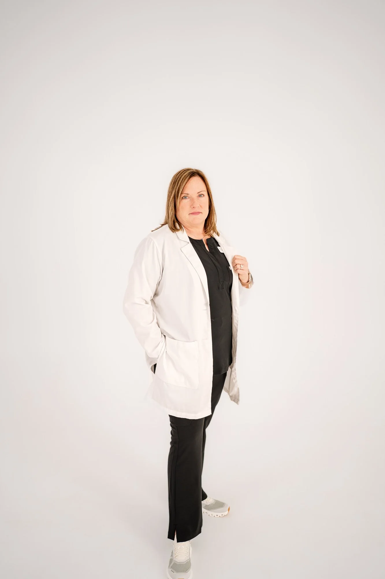 A woman with shoulder-length brown hair wearing a white lab coat over a black shirt, black pants, and white sneakers, standing against a plain light-colored background.