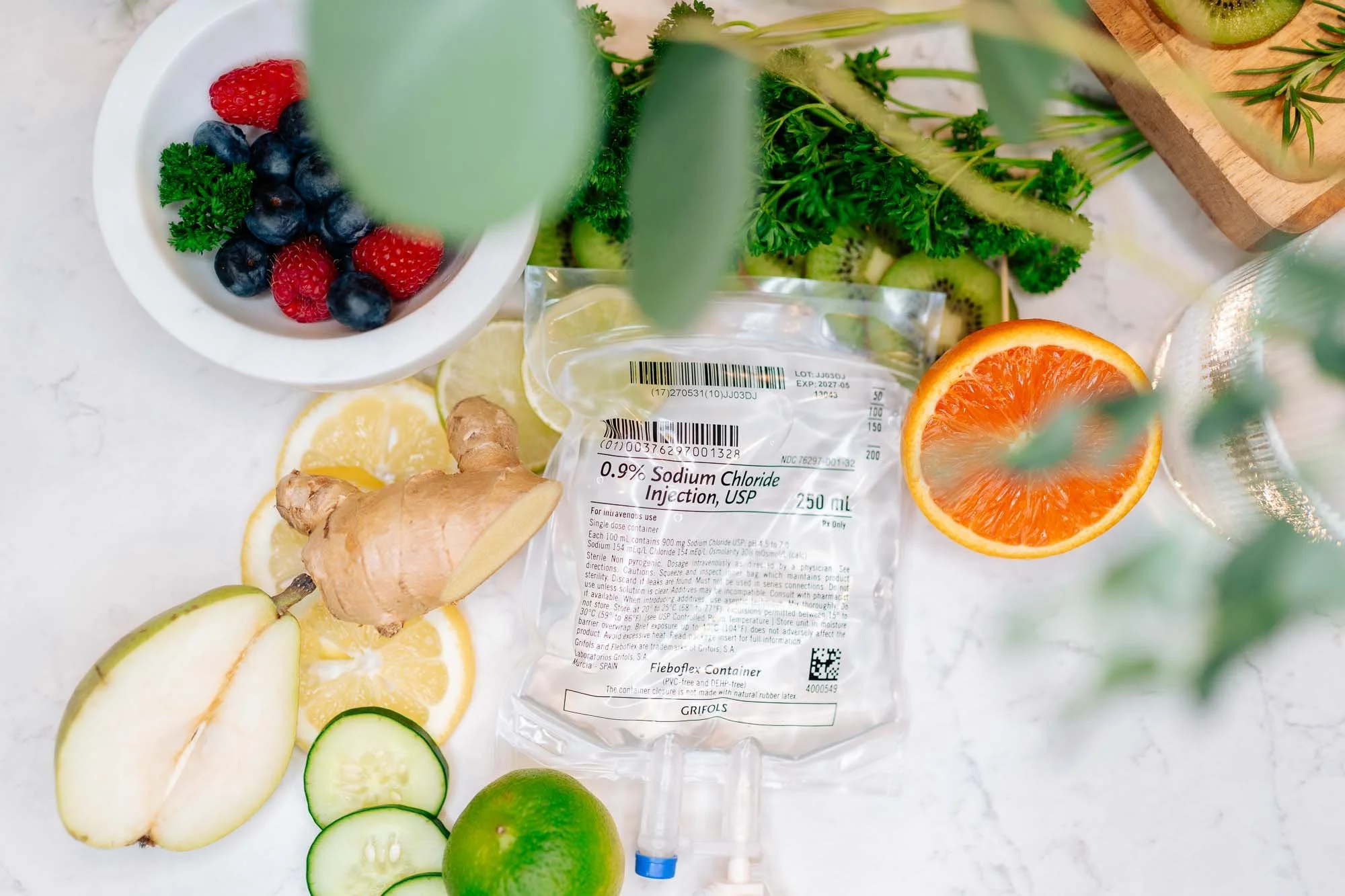 A variety of fresh fruits including blueberries, strawberries, lemon slices, cucumber slices, a lime, and half of an orange. Also, there is a packet of sodium chloride injection and a small bunch of parsley.