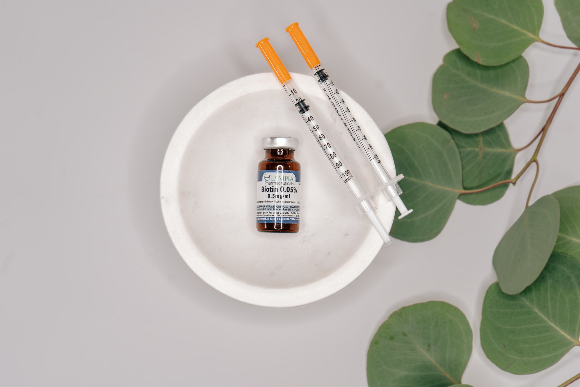 A small brown medicine bottle labeled 'Biotin 0.05%' with a white cap, two syringes with orange needles, all placed on a white dish, with green eucalyptus leaves nearby.