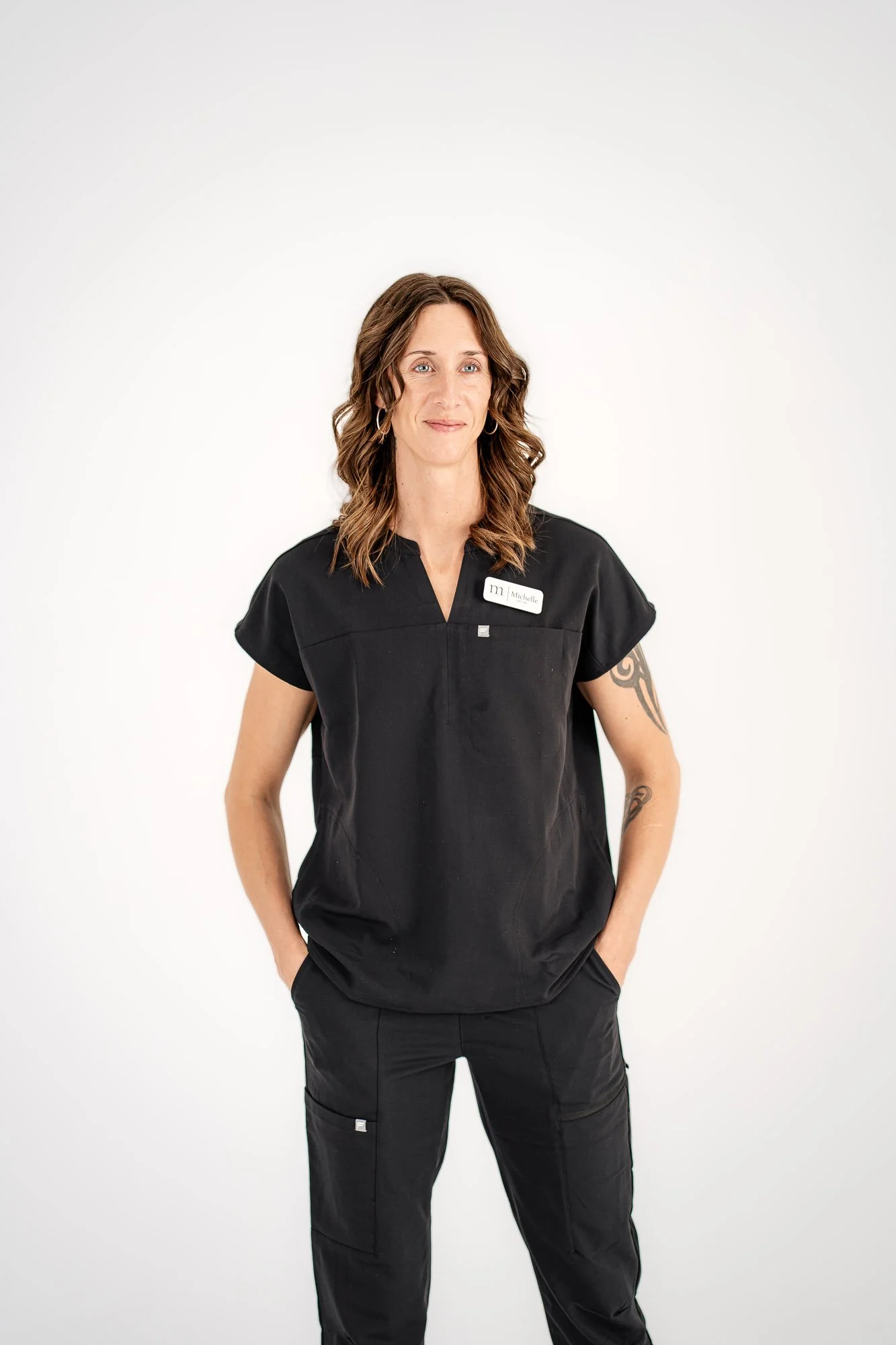 A woman wearing black scrubs with a name tag that says Michelle, standing with her hands in her pockets against a plain white background.