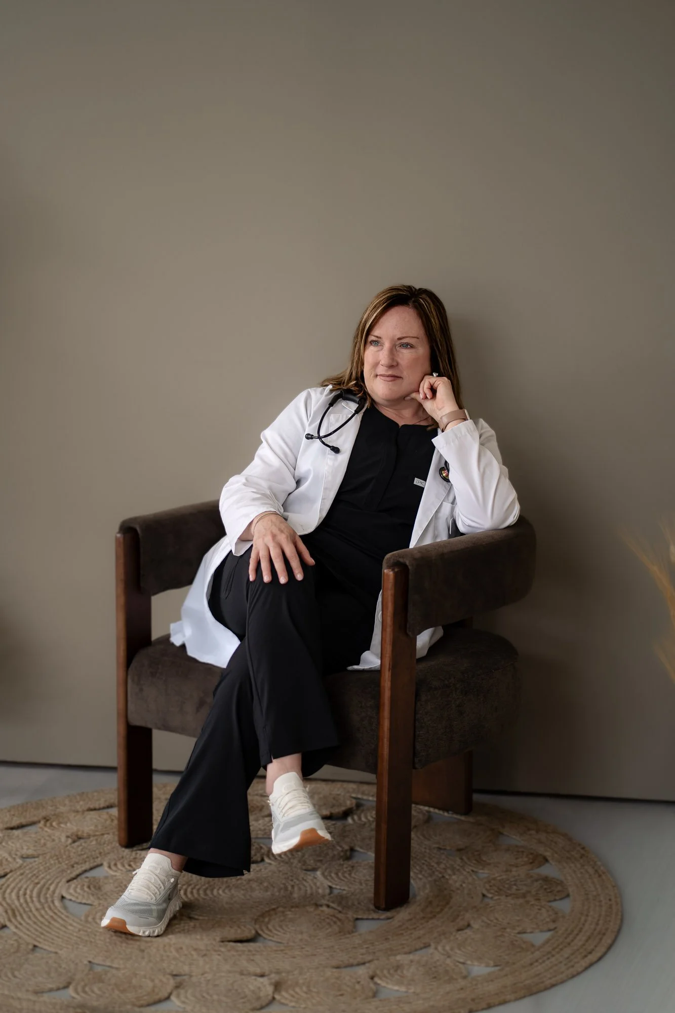 A woman in medical scrubs and a white medical coat sitting on a wooden chair with a dark cushioned seat in a room with a plain beige wall. She has a stethoscope around her neck and is wearing white sneakers. She has brown hair and is resting her chin on her right hand, looking contemplative.