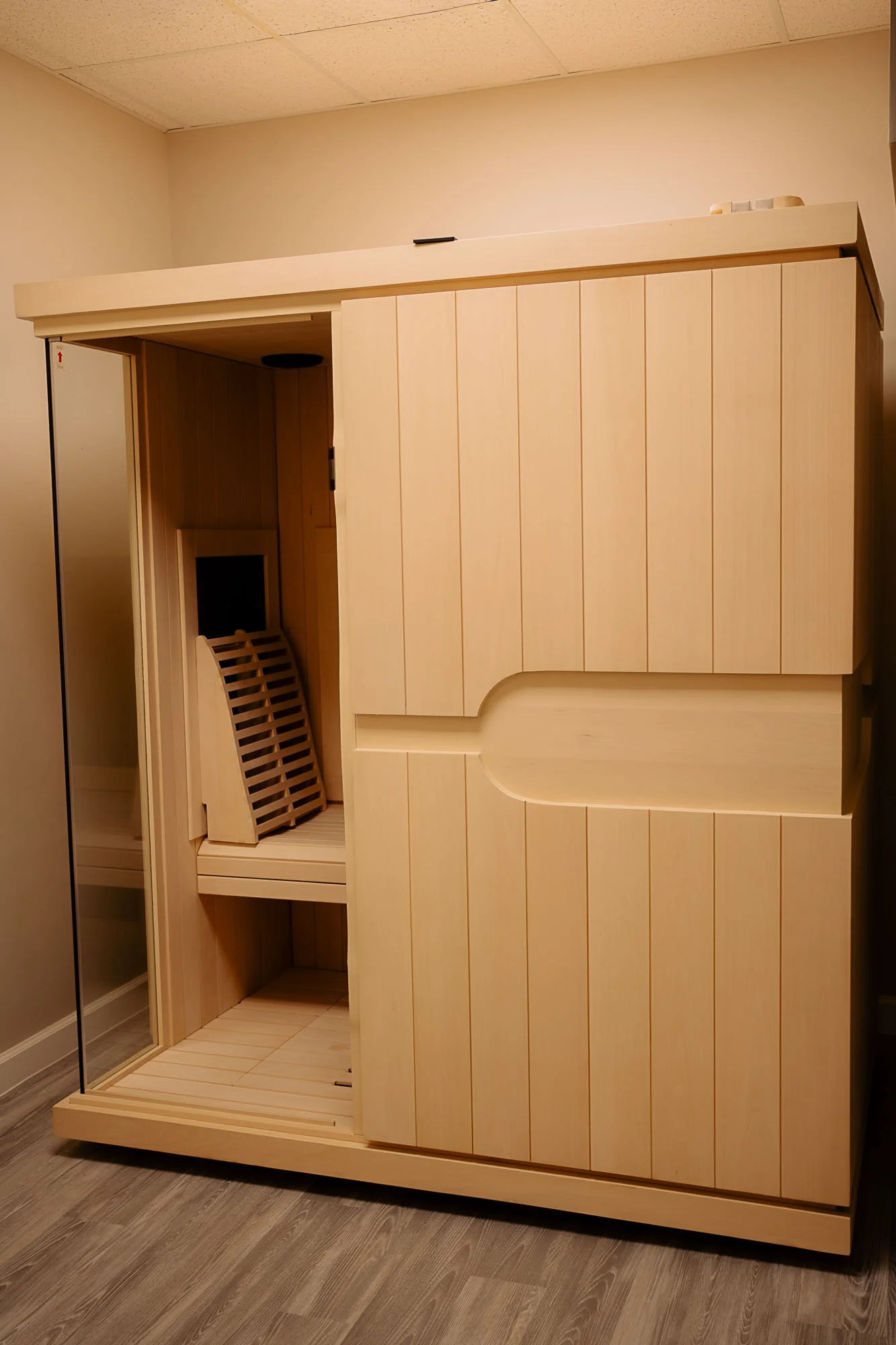 A large wooden sauna with a glass door on the left, and a built-in bench inside. The sauna has a light wood finish and is situated on a wooden floor.