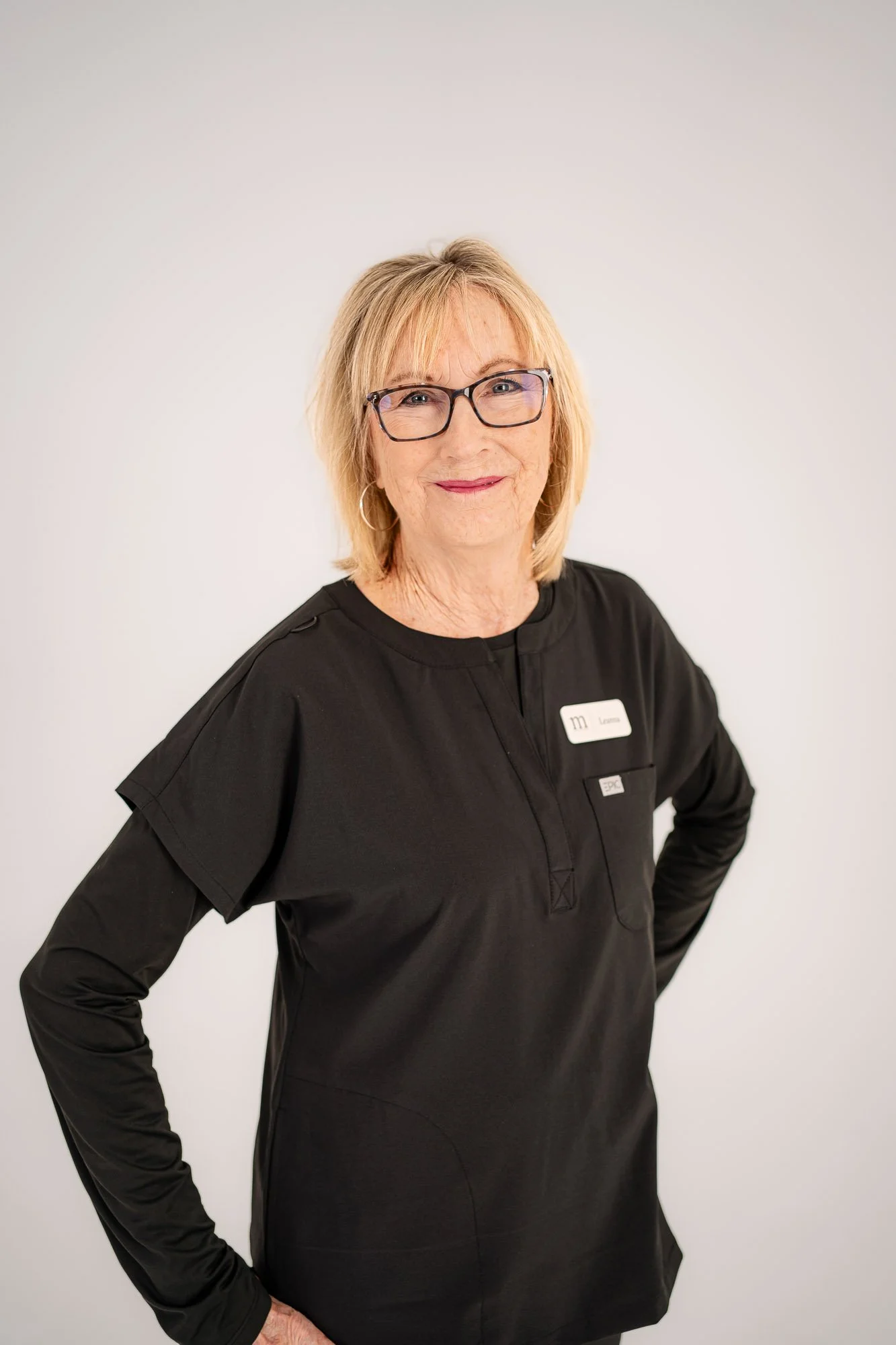 A smiling middle-aged woman with blonde hair, glasses, and hoop earrings wearing a black uniform with a name tag, standing against a plain white background.