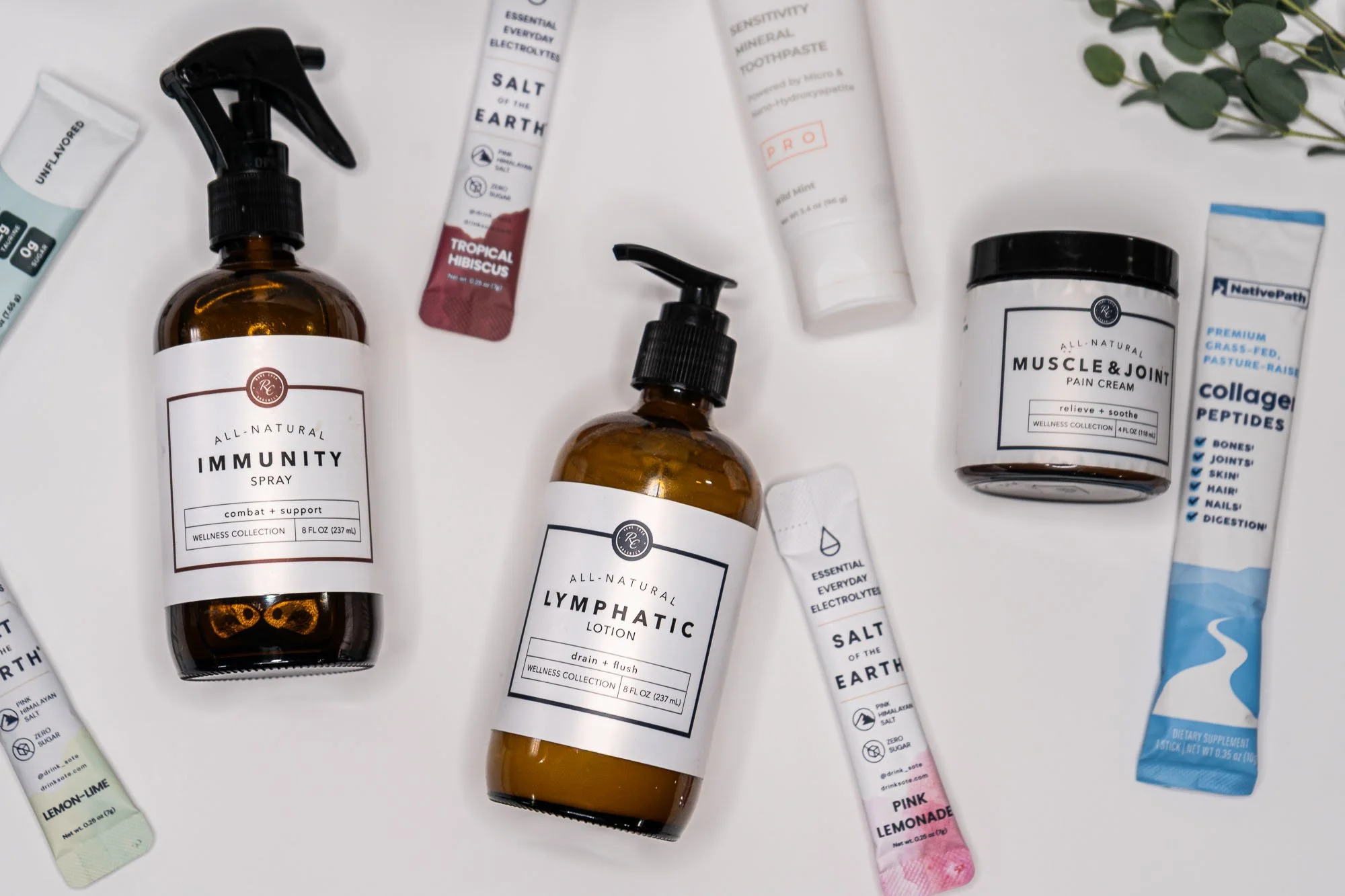 Assortment of natural wellness products including spray bottles labeled immunity and lymphatic, a muscle and joint pain balm, toothpaste, and supplements, along with eucalyptus leaves.