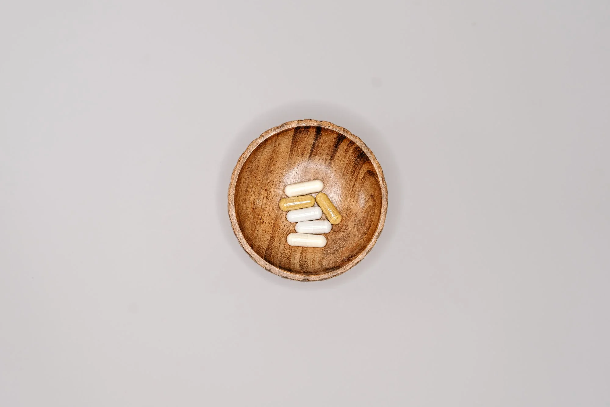 A small round wooden bowl containing white and yellow capsules placed on a white surface