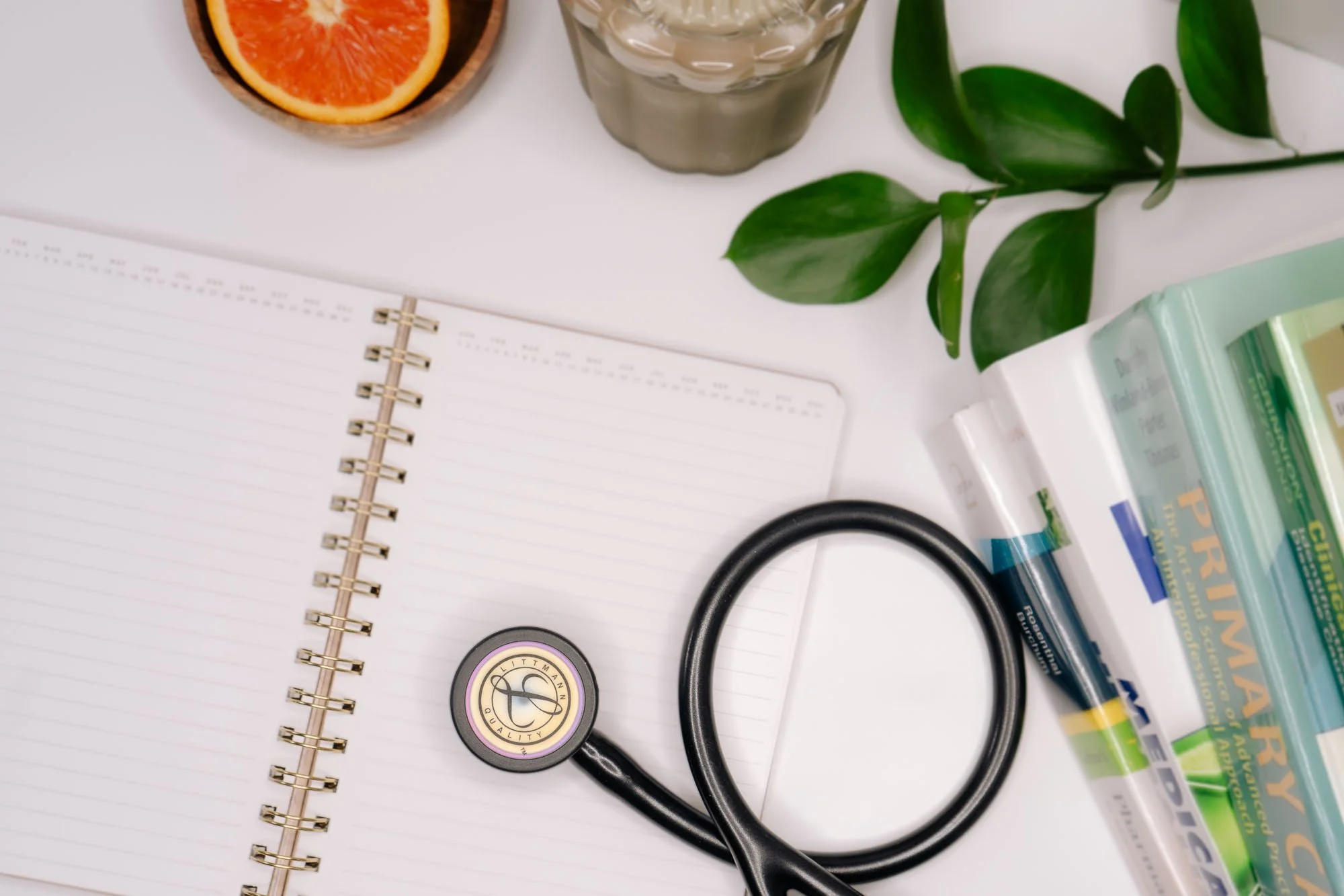 A flat lay of medical and health-related items on a white surface, including a stethoscope, a notebook, a glass of water, a halved grapefruit, green leaves, and boxes of medication or supplements.
