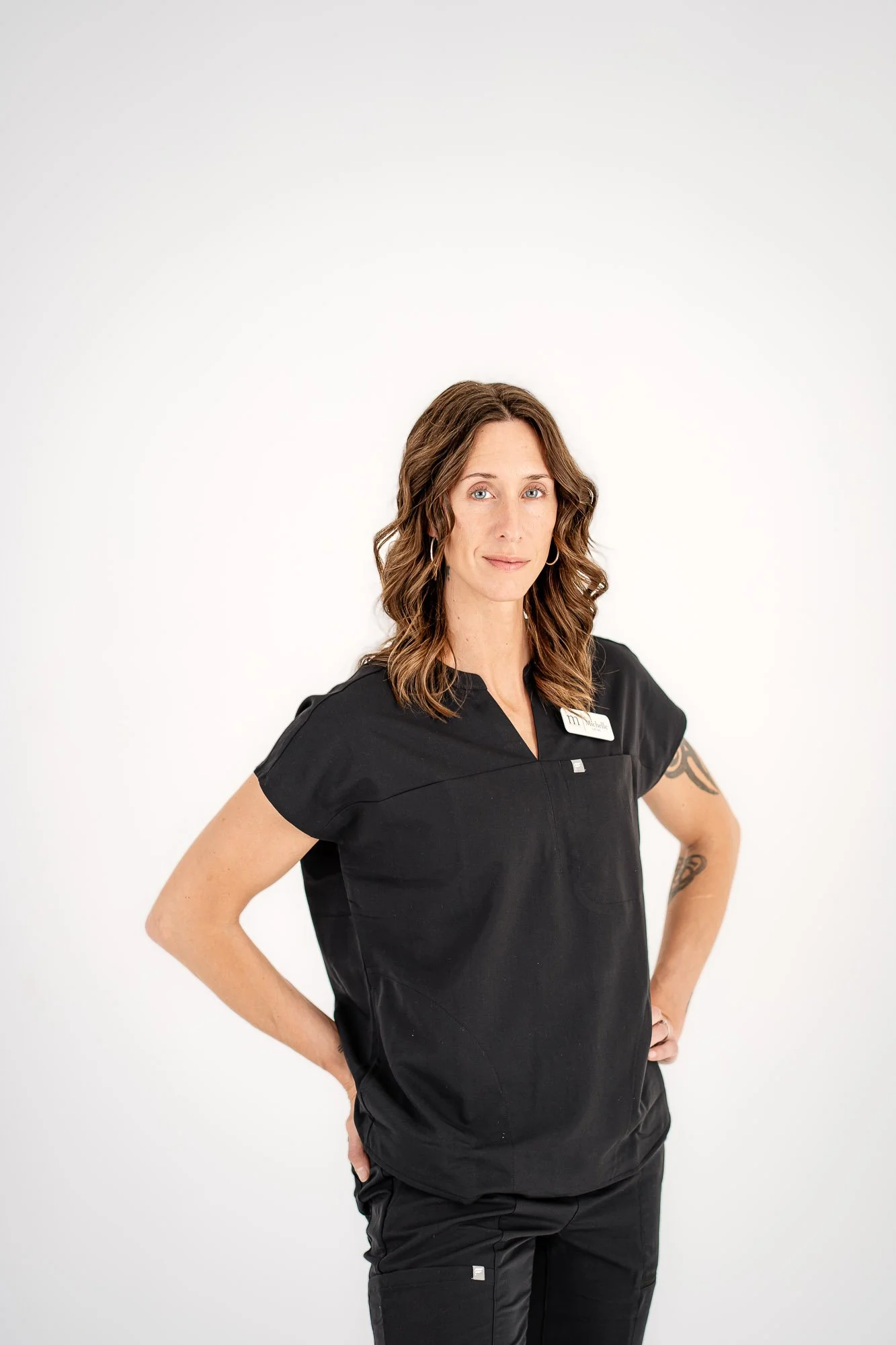A woman with wavy, shoulder-length brown hair and blue eyes wearing a black medical uniform with a name tag and tattoo on her left arm, standing confidently with hands on hips against a plain white background.