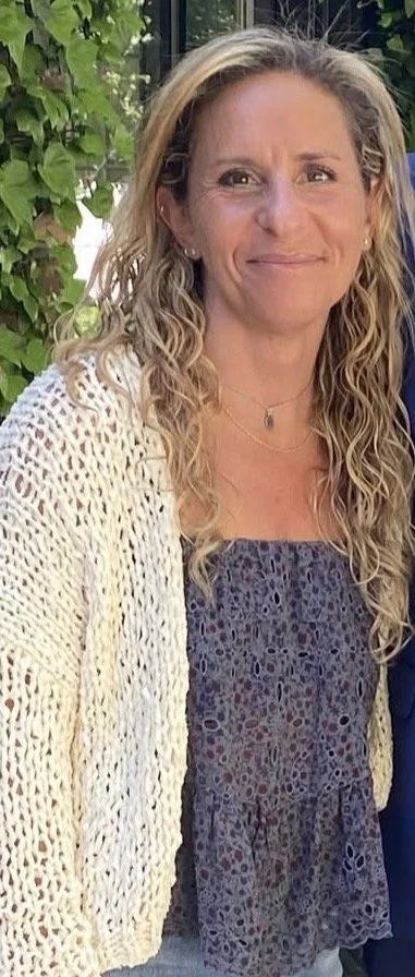 A woman with blonde, curly hair smiling outdoors, wearing a light-colored cardigan over a patterned dress, with green foliage in the background.