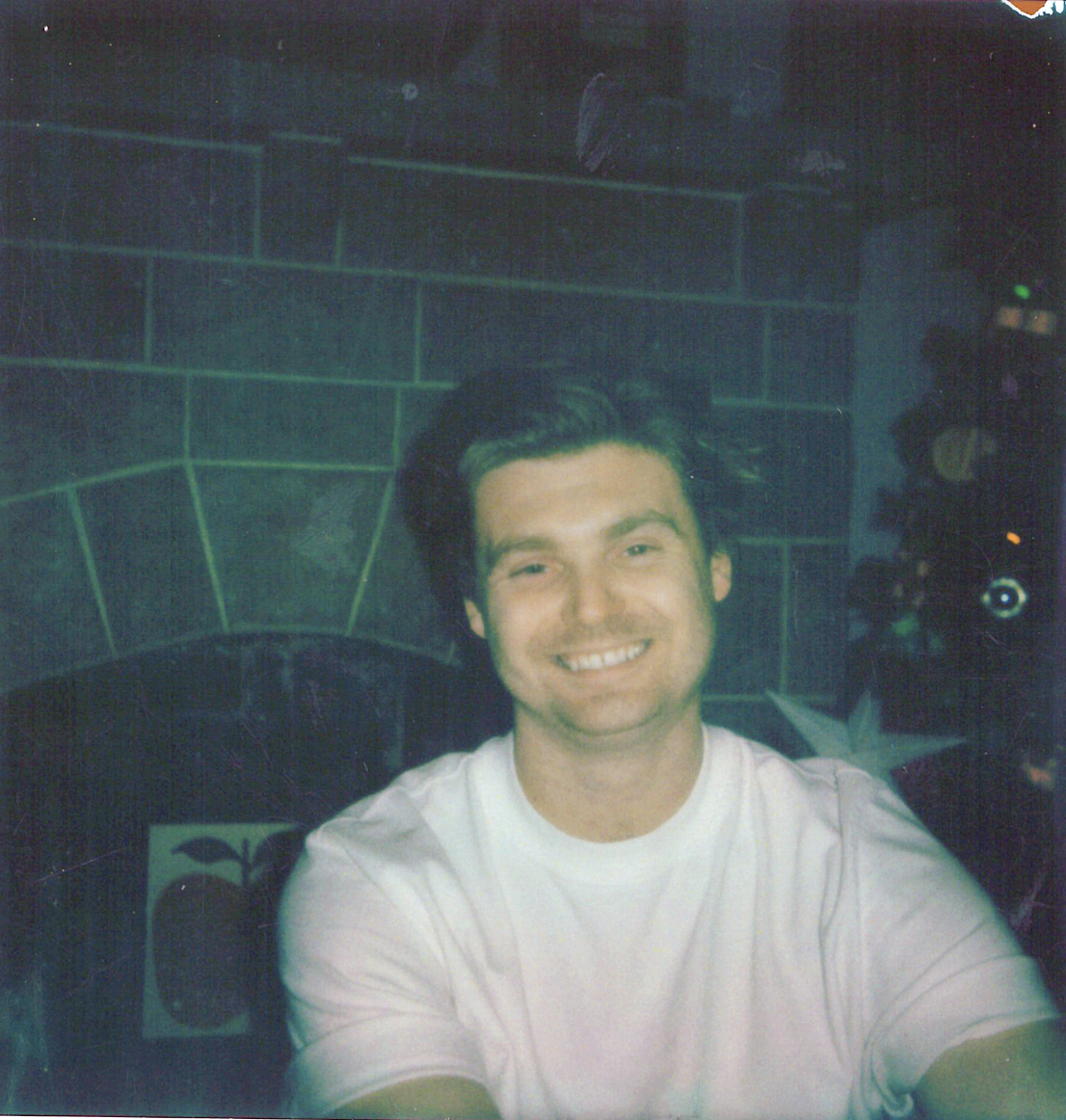 A young man with dark hair smiling, wearing a white t-shirt, sitting in front of a dark green tiled wall with a fireplace and some lights and objects in the background.