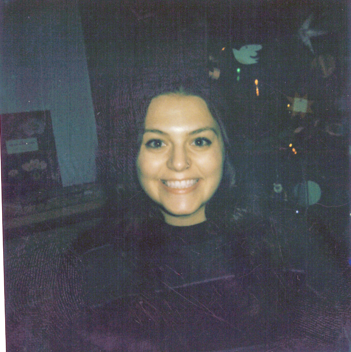 Close-up of a young woman with short dark hair, smiling, in a dark indoor setting with neon lights and a Twitter logo in the background.