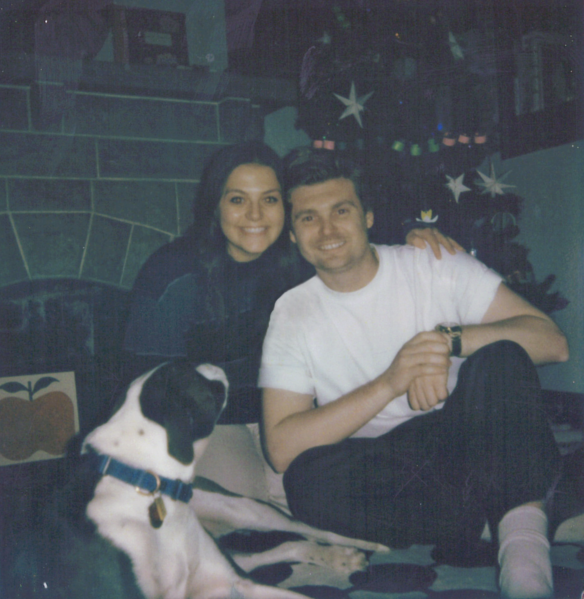 Two smiling young adults, a woman and a man, sitting together with a black and white dog in front of a decorated Christmas tree. The woman has dark hair, and the man has short dark hair, wearing a white T-shirt. The background features a Christmas tree with star ornaments and decorations, and a painting or picture on the wall. The dog is sitting on the floor, looking up, with a collar and tags.