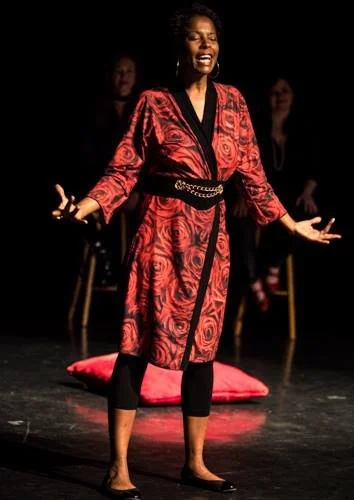 Woman performing on stage wearing a red and black patterned robe, with two women sitting in the background.