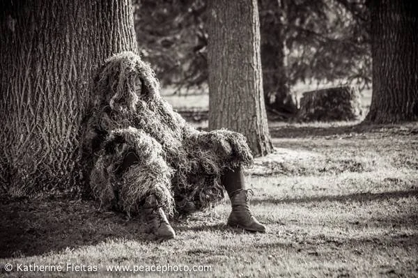 Person sitting against a tree in a camouflage ghillie suit with boots, in a wooded area.