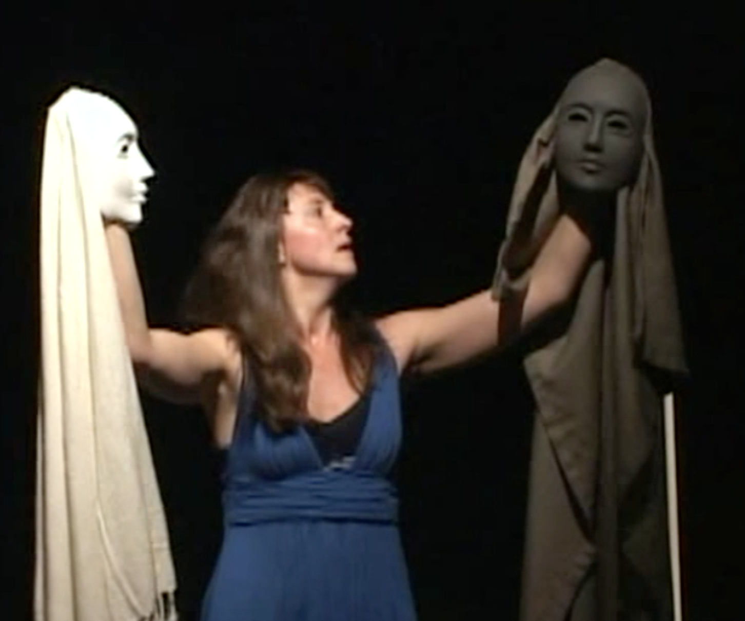 A woman in a blue dress interacts with two mannequins of women’s faces draped in scarves against a black background.