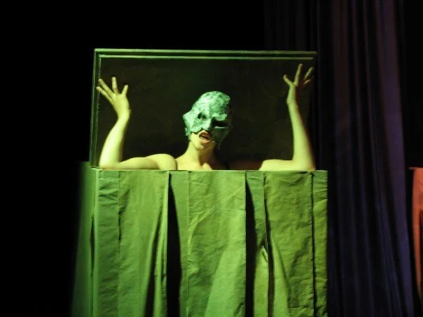 Performer wearing a mask and costume behind a green theater prop with a curtain, on stage.