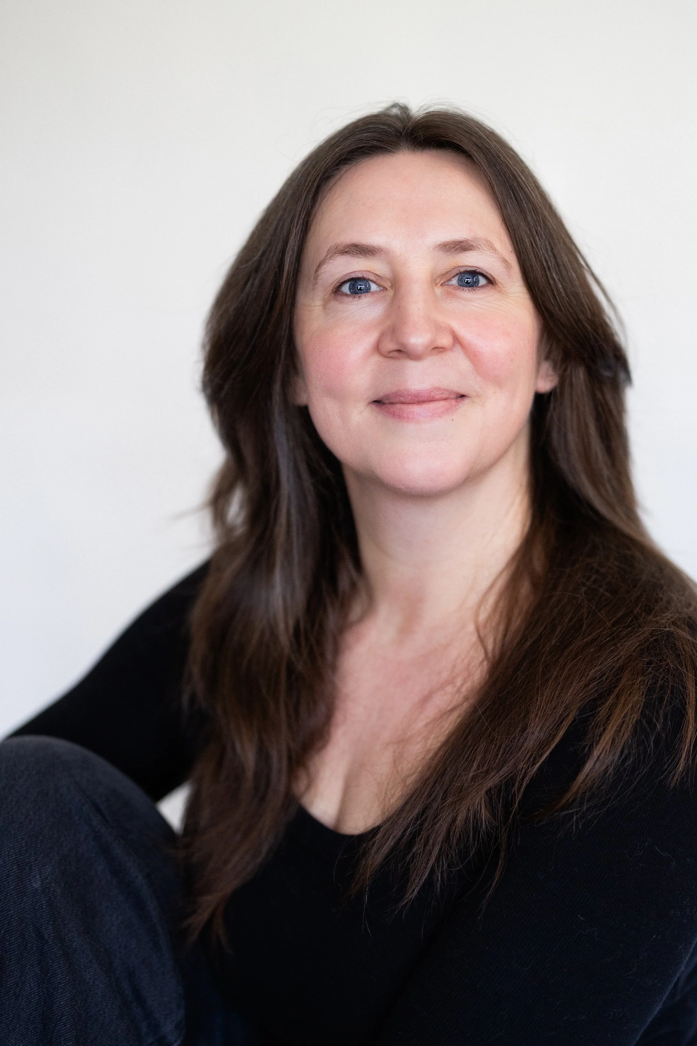 Close-up portrait of a woman with long brown hair, blue eyes, and fair skin, smiling softly, wearing a black top, against a plain white background.