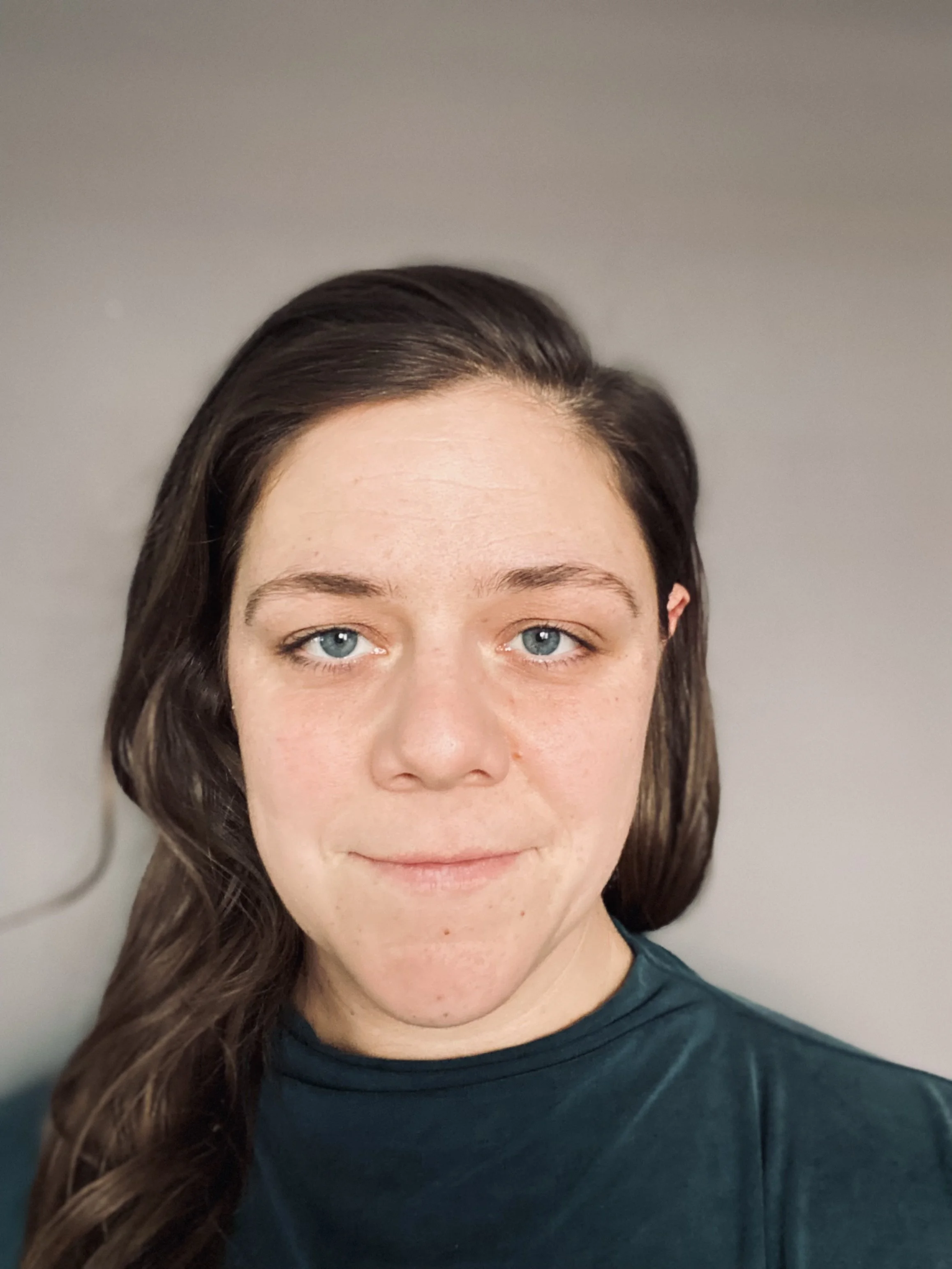 Close-up of a woman with long brown hair and blue eyes, wearing a dark teal shirt, against a plain background.