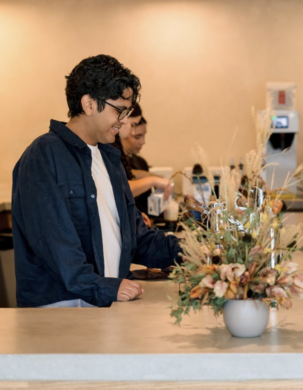 A young man smiling at a floral display behind a counter in a floral shop, with two other people in the background working.