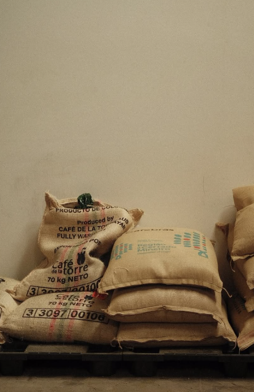 Stacks of burlap sacks on a pallet, with some partially hidden behind others, on a warehouse floor against a plain wall.