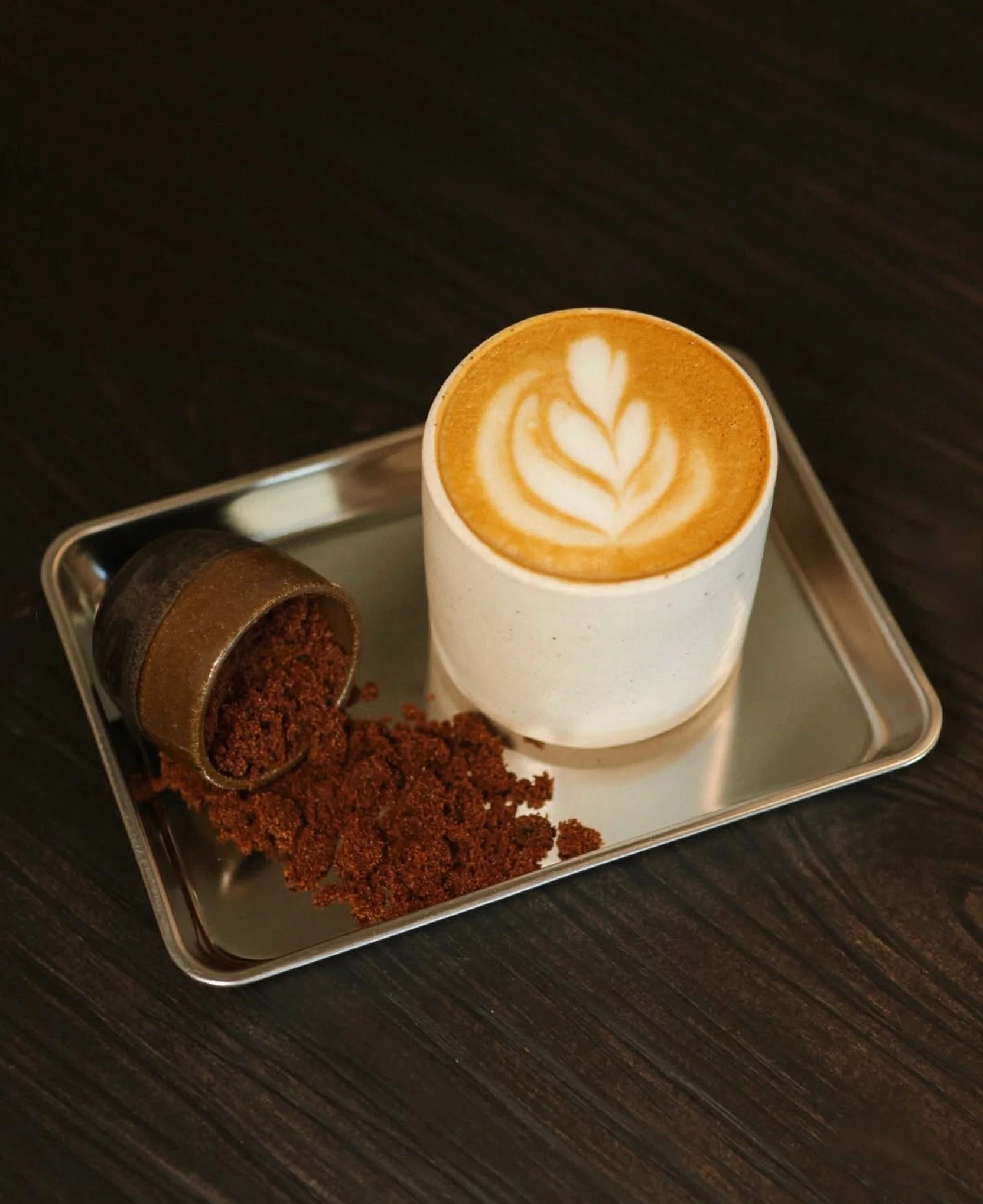 A latte with latte art in a white cup on a metal tray, with coffee grounds spilled from a small metal container next to it, on a dark wooden table.