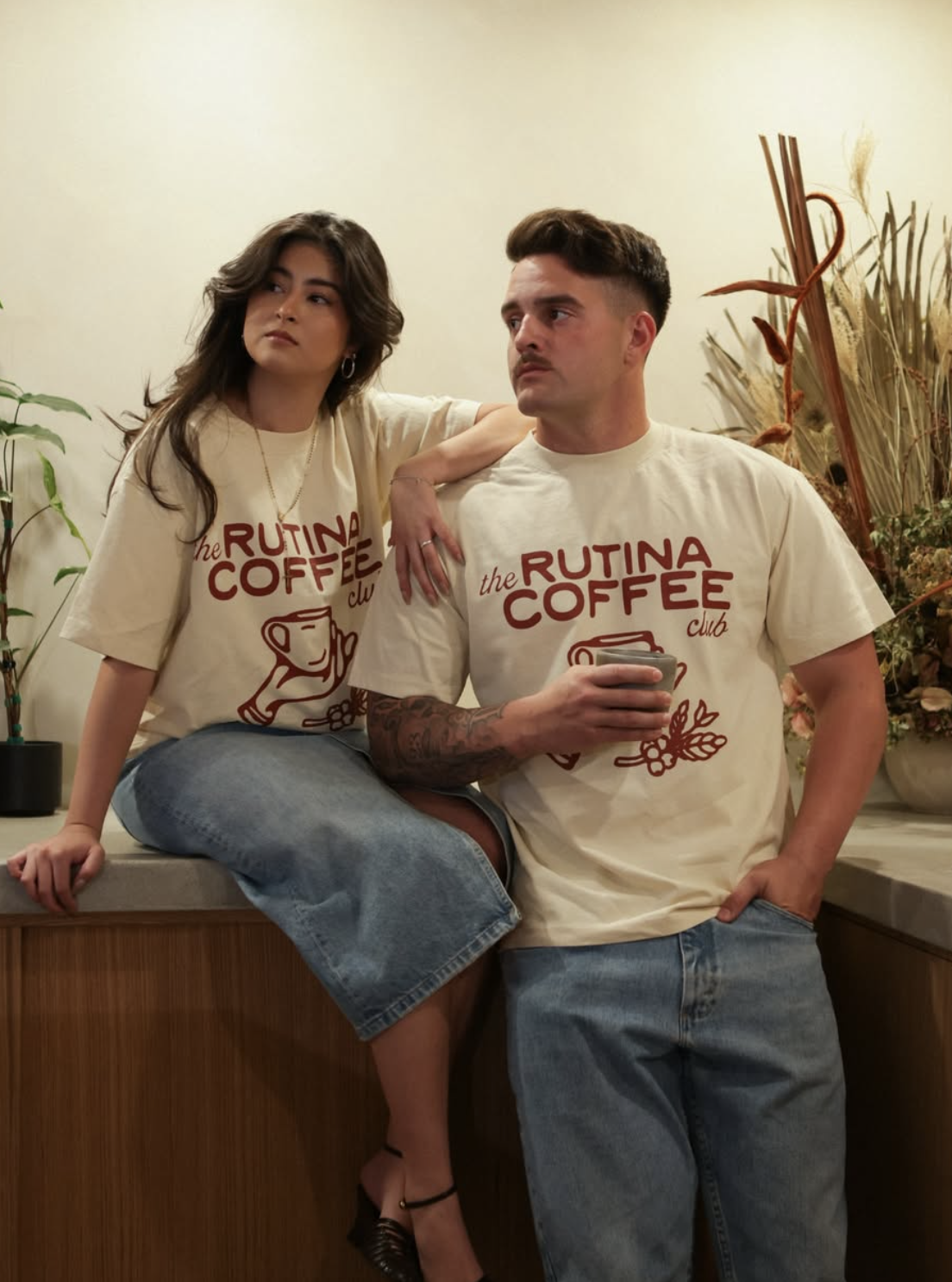 Two young adults, a woman sitting on a counter with her arm around a man, both wearing cream-colored T-shirts with 'the RUTINA COFFEE club' logo. The woman has dark wavy hair, and the man has short dark hair and a tattooed arm, holding a coffee cup. They are in a cafe with plants and decorative dried flowers in the background.