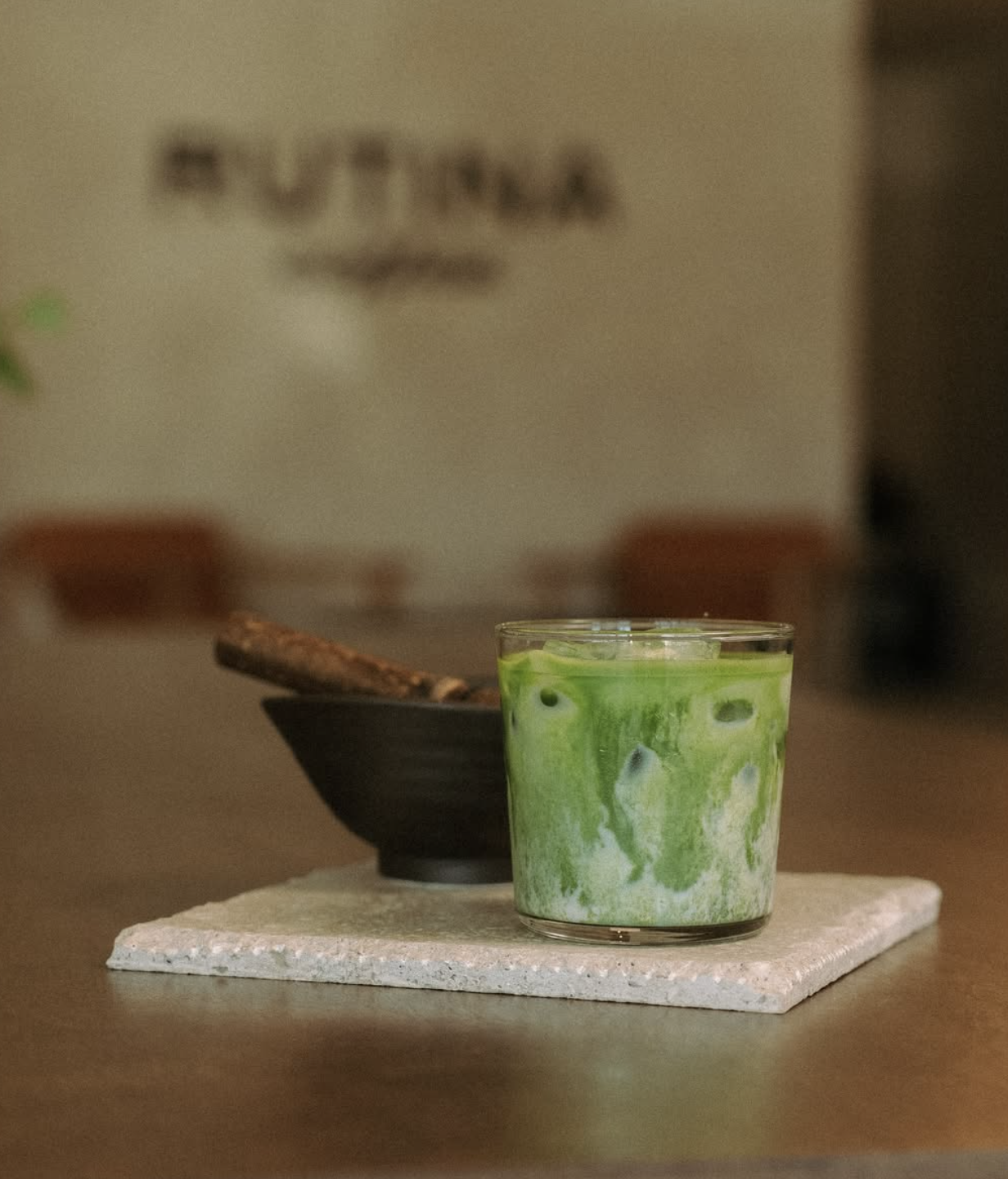 A glass of green tea with ice on a table, with a black bowl and a paper napkin in the background.