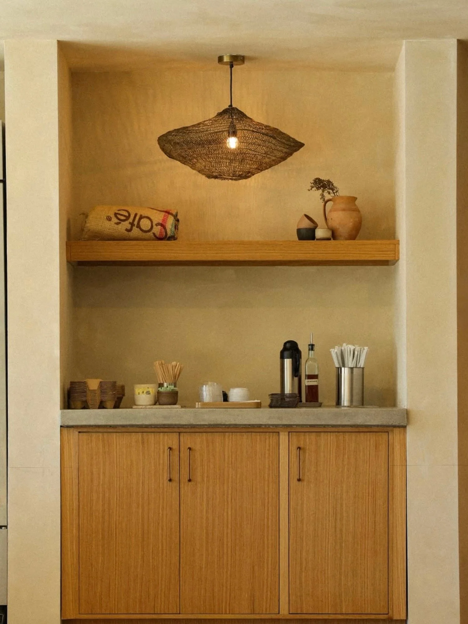 A small kitchenette with wooden cabinets, a countertop with coffee and tea supplies, and a shelving area above with decorative items and a woven pendant light.