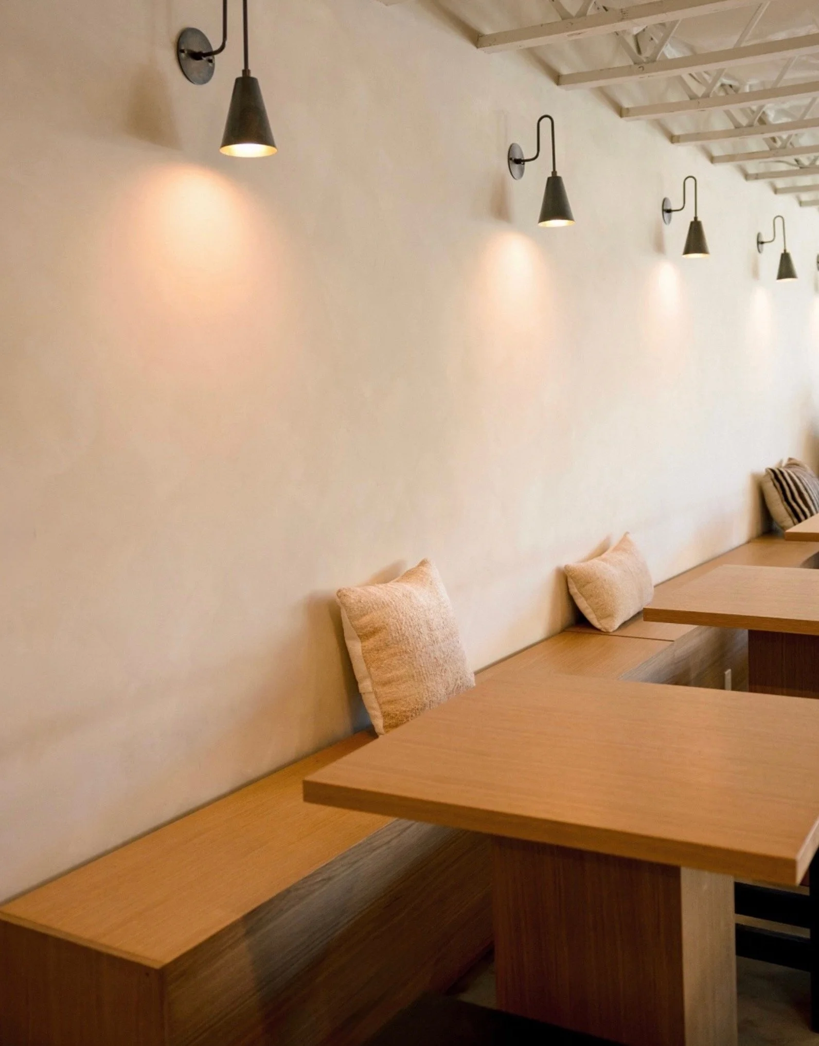 A row of small wall-mounted black lamps casting light onto a plain beige wall, with a wooden bench and pillows below.